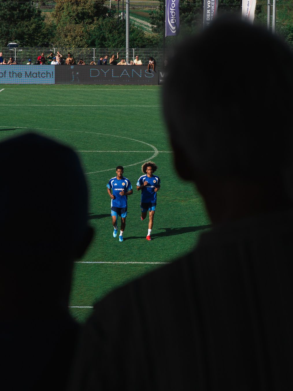 Two football players in blue jerseys jog on a green pitch, viewed from behind silhouetted spectators.