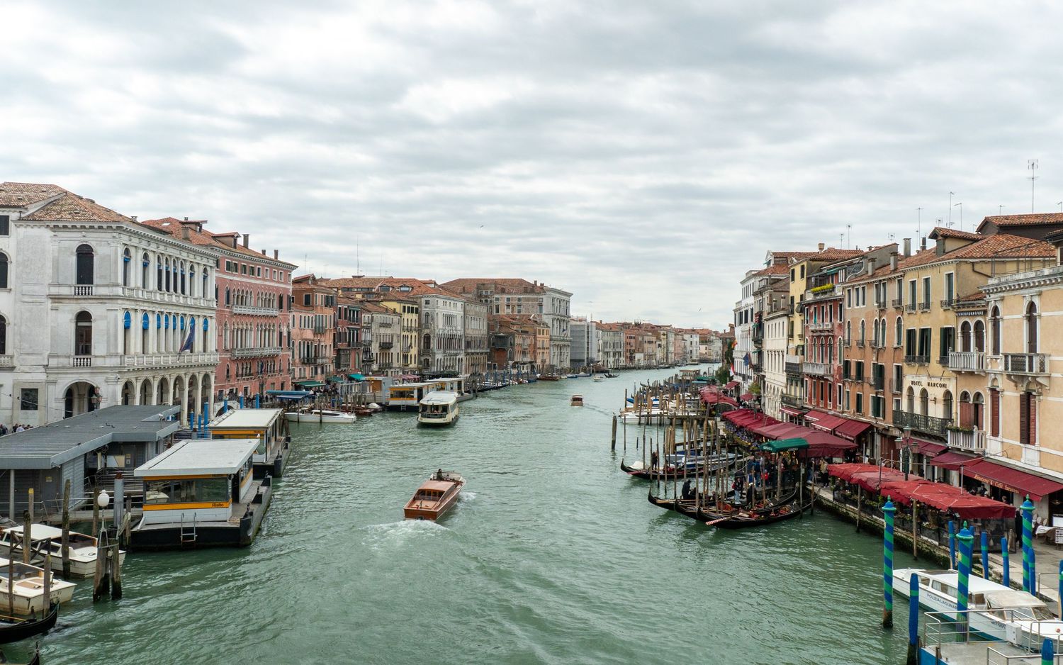 Aerial view of Venice Grand Canal with gondolas, boats, and colorful historic buildings lining the waterway under cloudy skies.