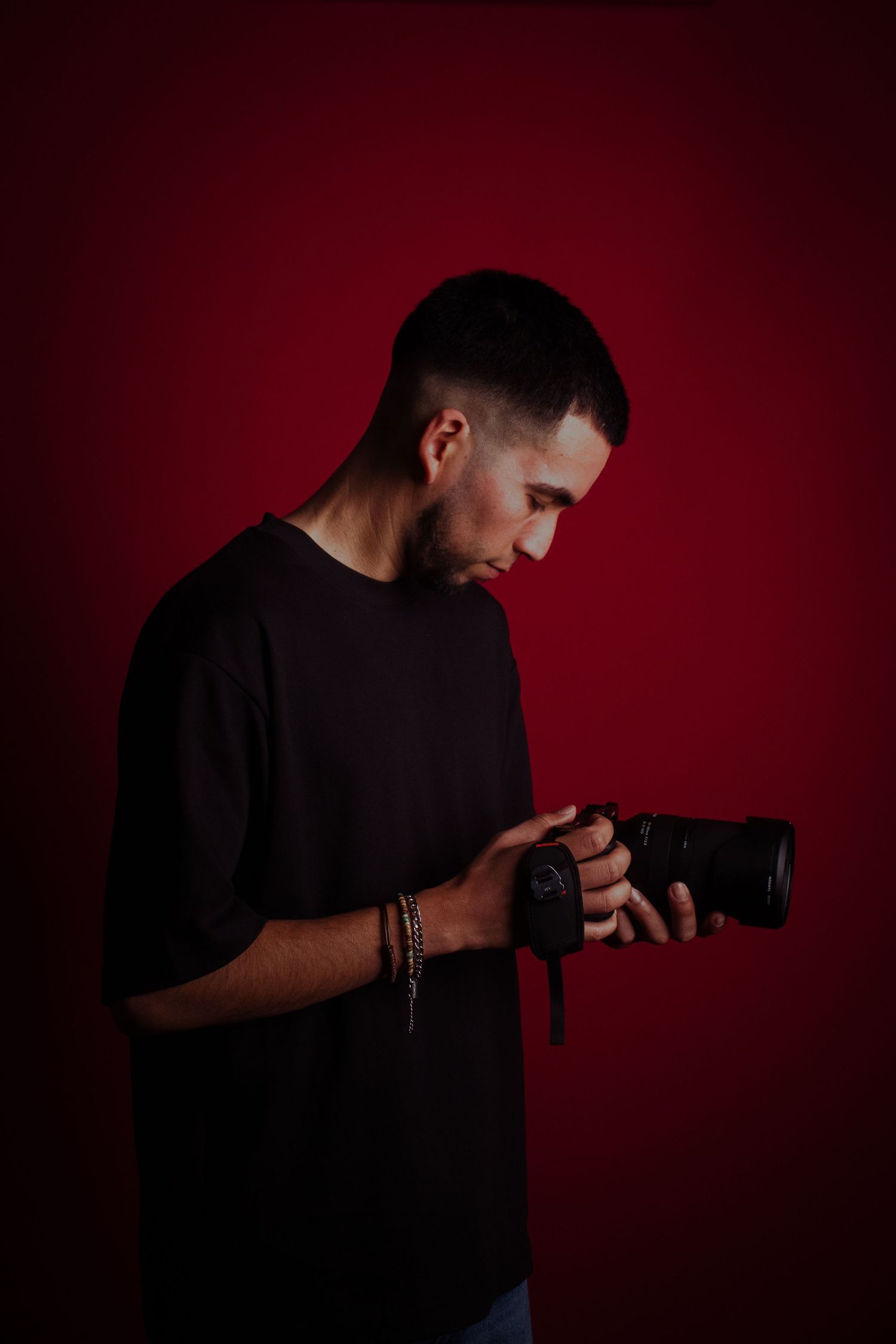 A photographer in a black shirt checks settings on their camera against a dark red background.