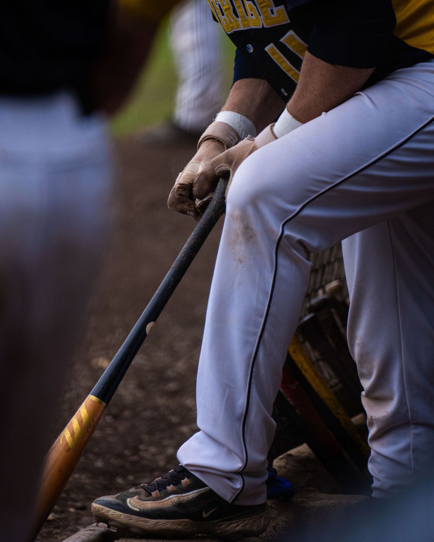 Baseball players in white uniforms sit in dugout during game while holding bats and wearing cleats.