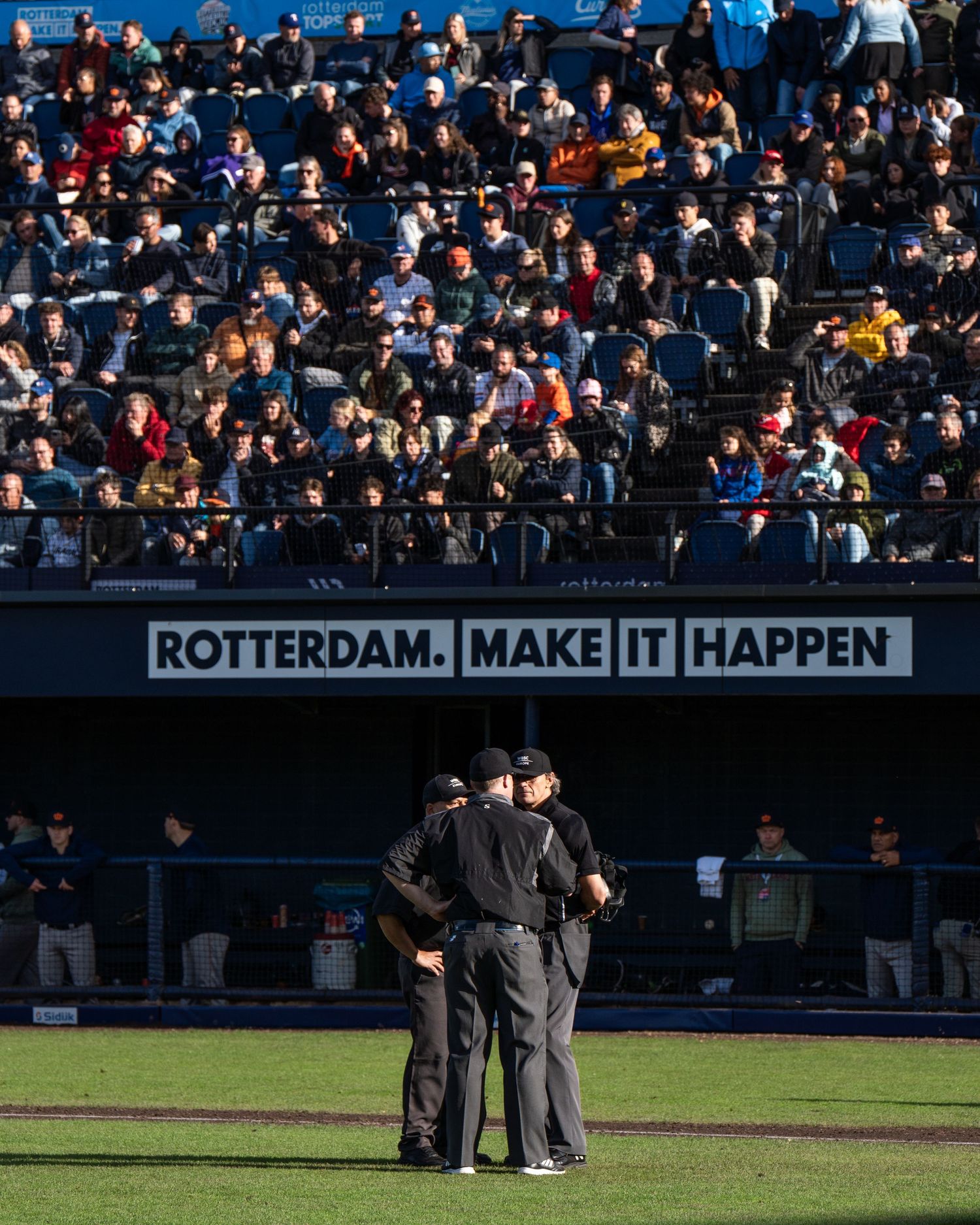 Baseball umpires gather on the field during a game with stadium crowd and signage visible in the background.