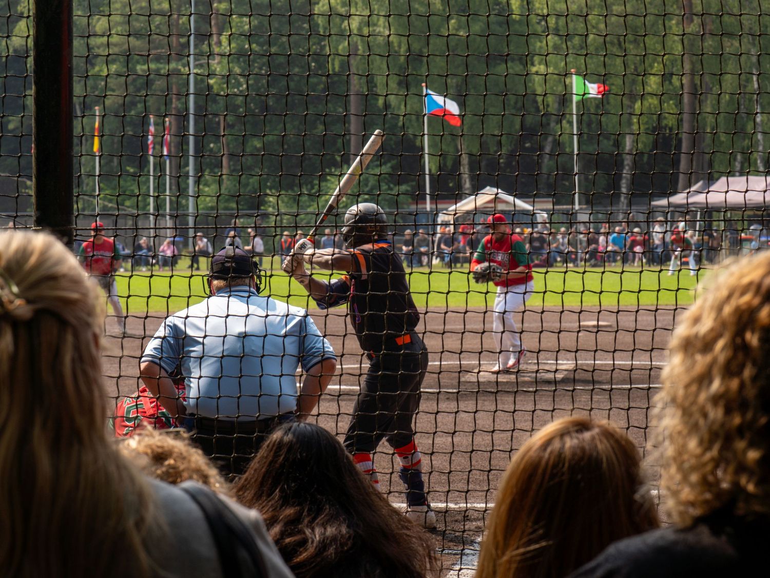 Spectators watch from behind a fence at an outdoor sporting event with flags waving in the background.