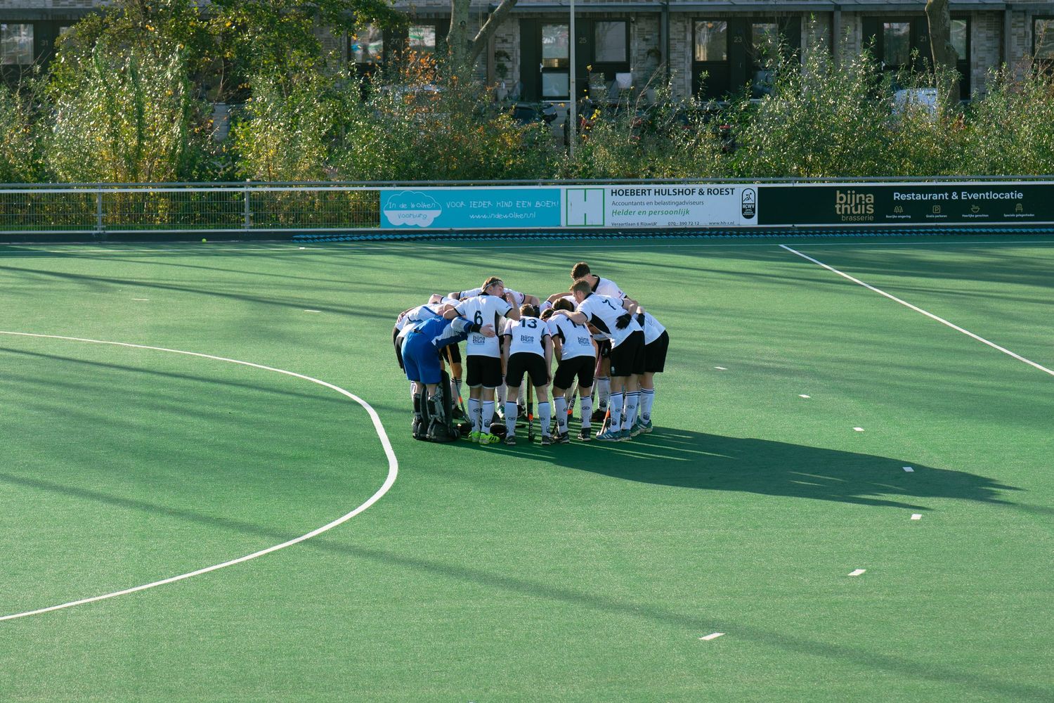 Soccer players gather in a huddle formation on a green artificial turf field during practice or pre-game preparation.