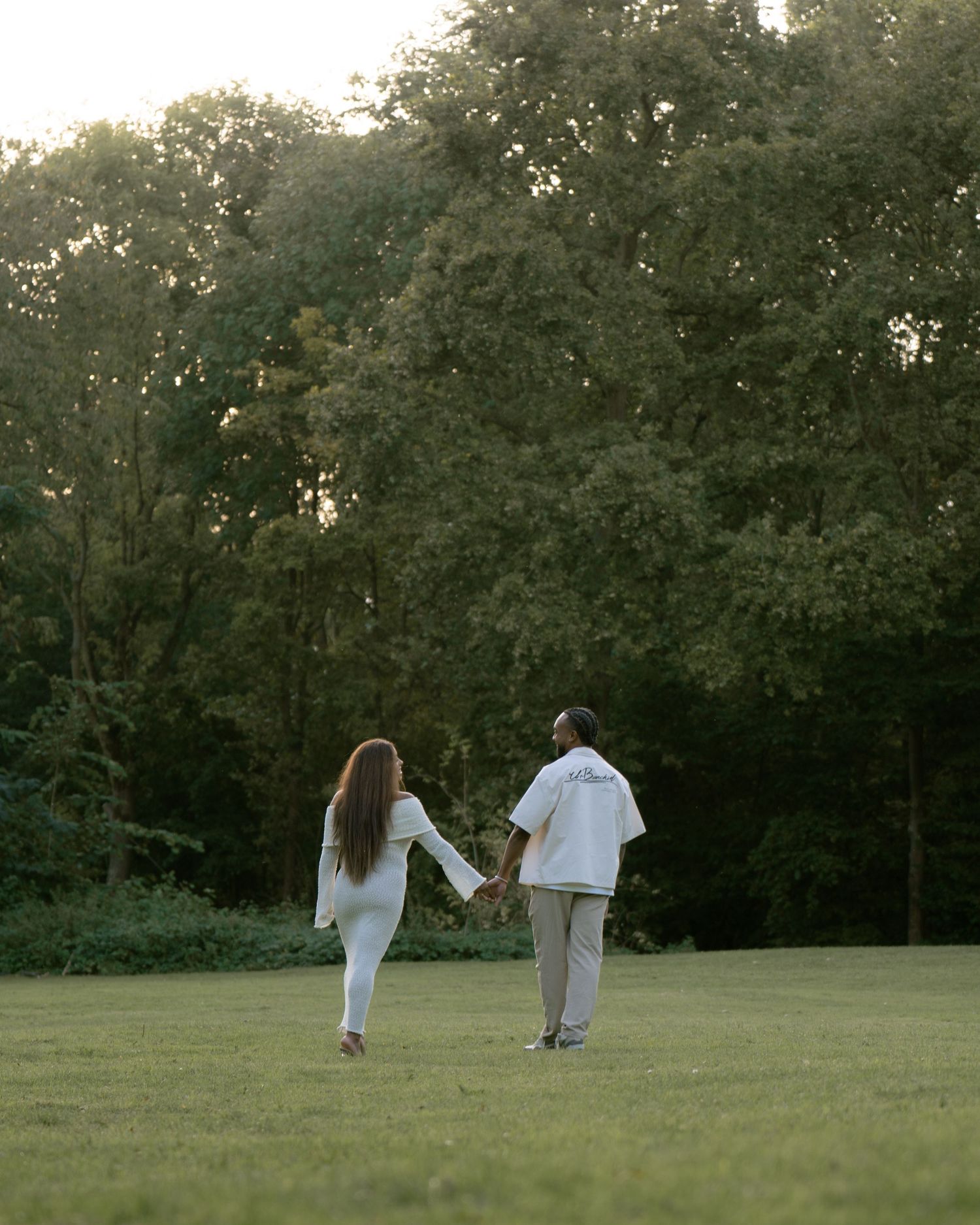 Two people in white clothing walk hand in hand across a serene grassy field at dusk with trees in the background.