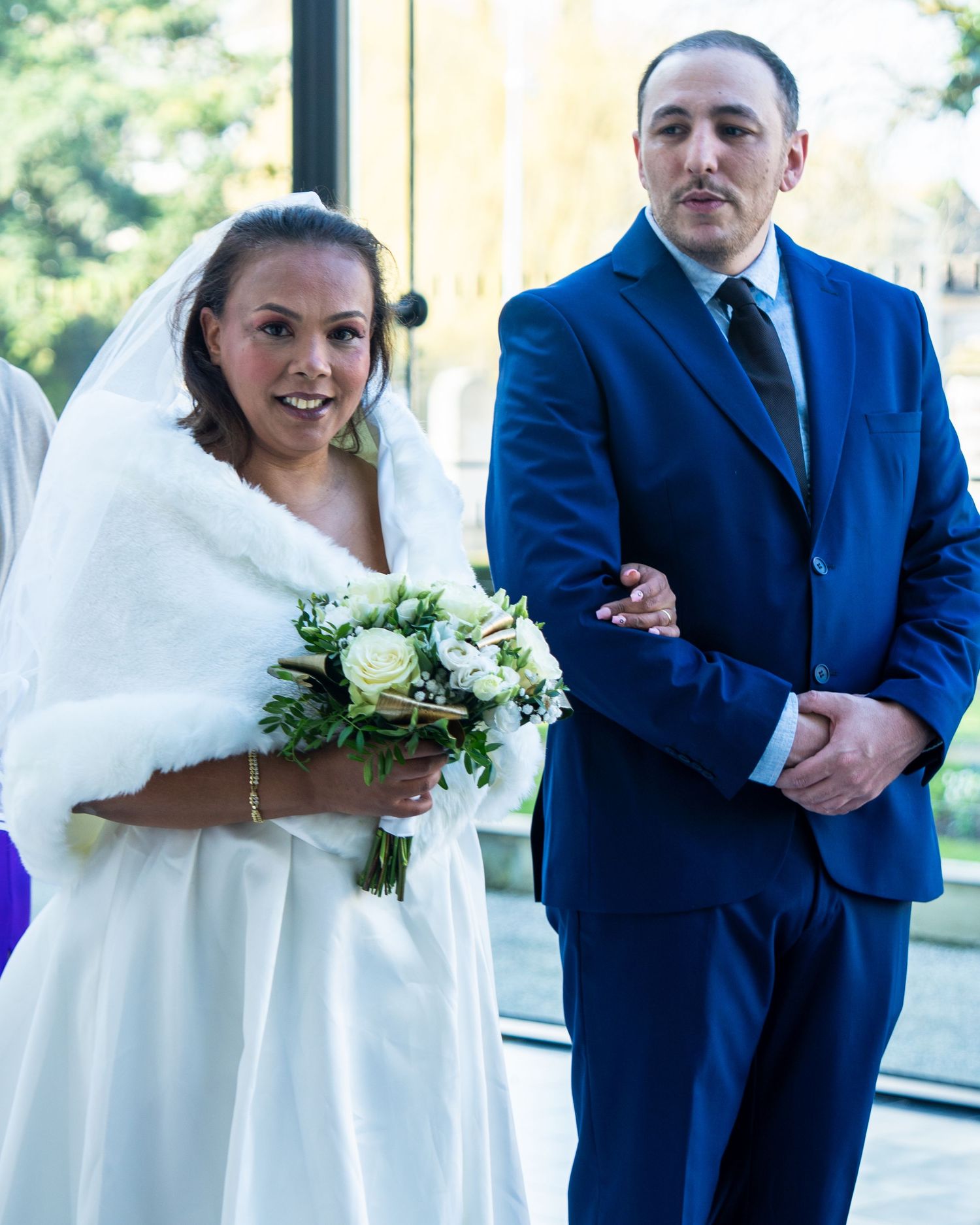 A bride in a white dress and fur shawl stands with a person in a blue suit holding a bouquet.
