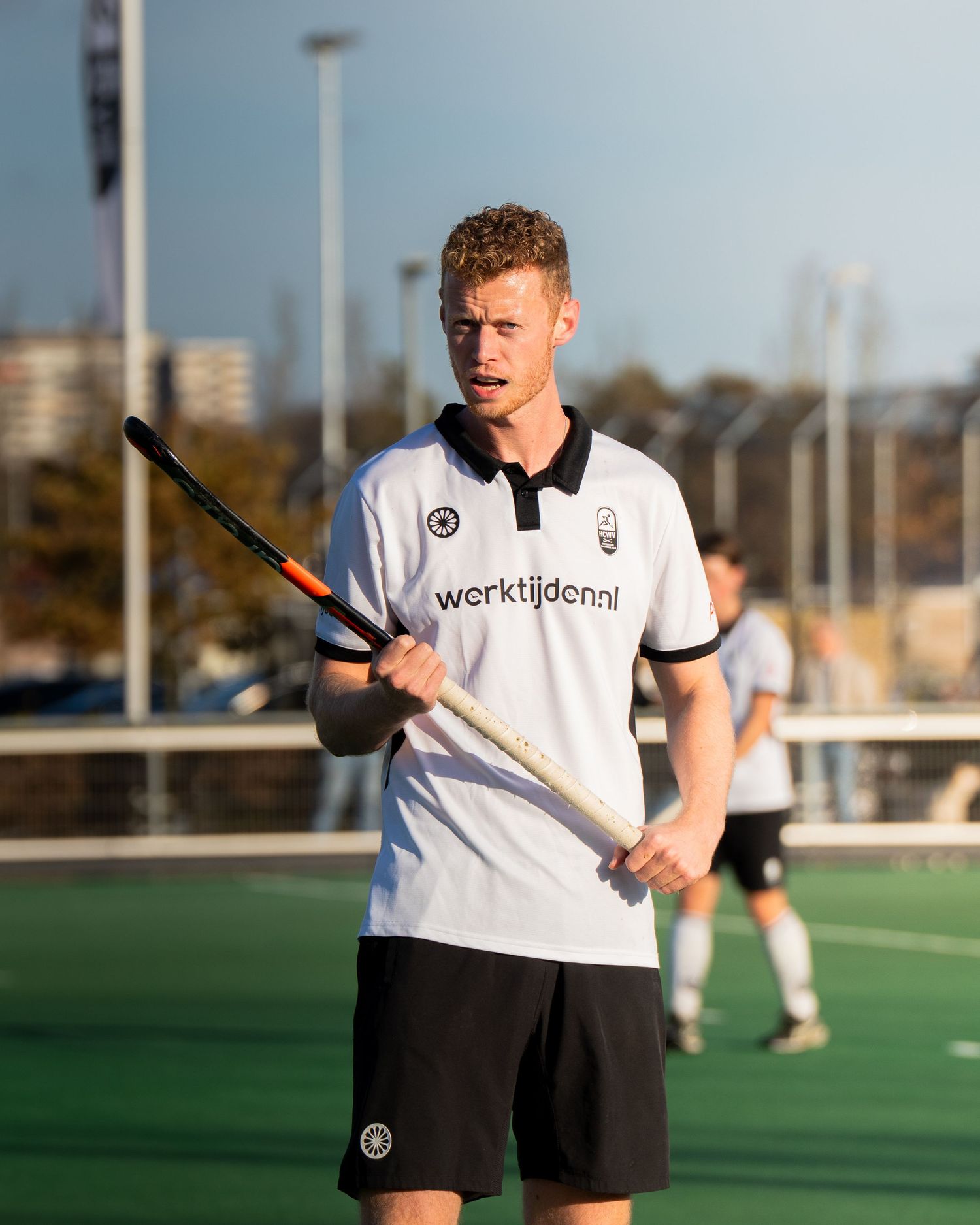 A hockey player in white and black uniform holds a stick on a green turf field during practice.