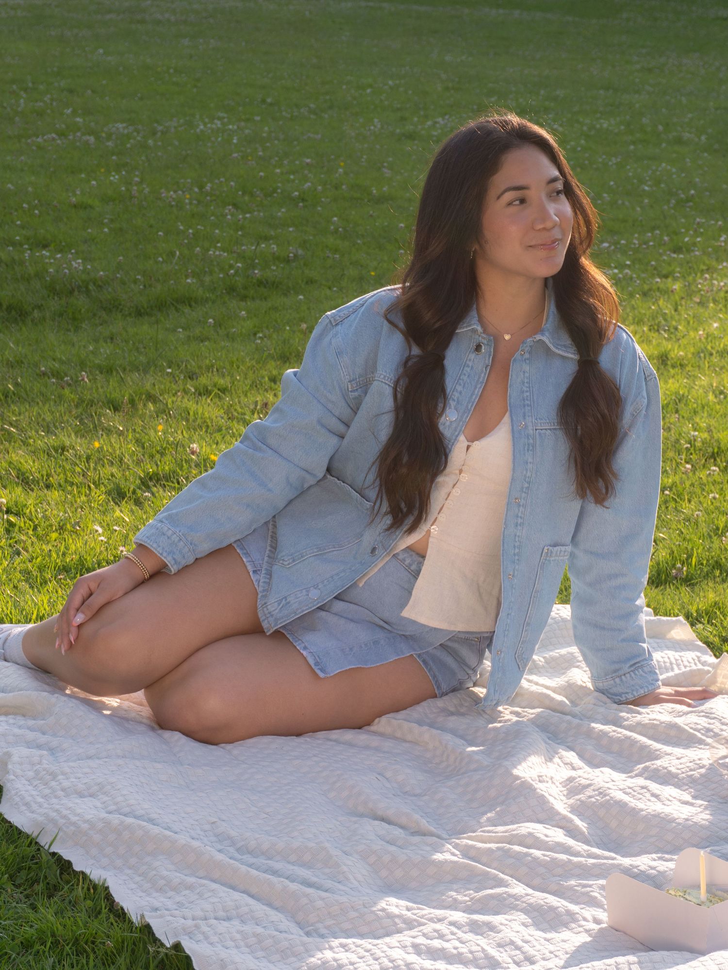 A person in a light denim jacket lounges on a white blanket in a sunny green field.