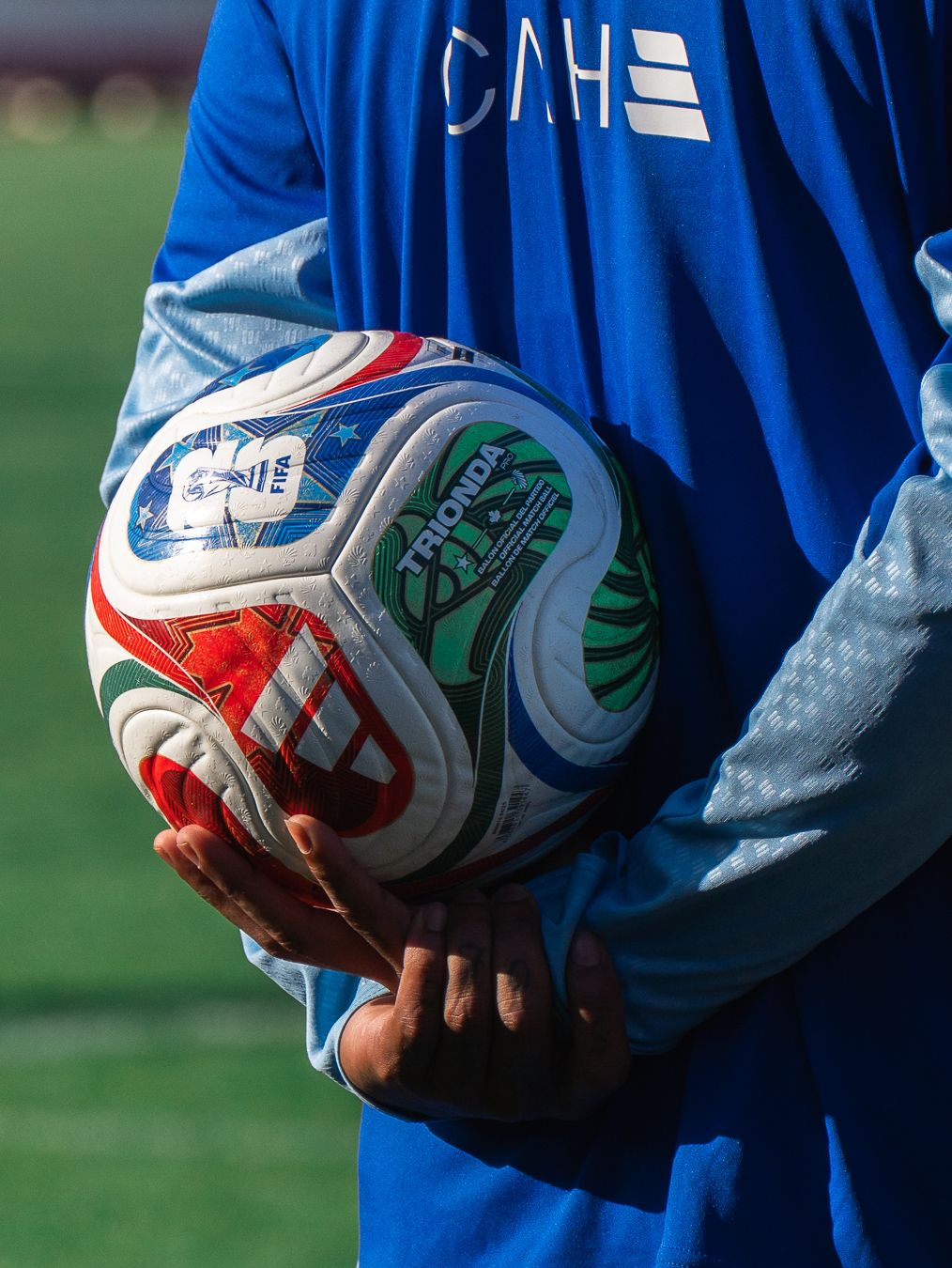 Soccer player in blue jersey holding a colorful Trondi match ball on a sunny field.