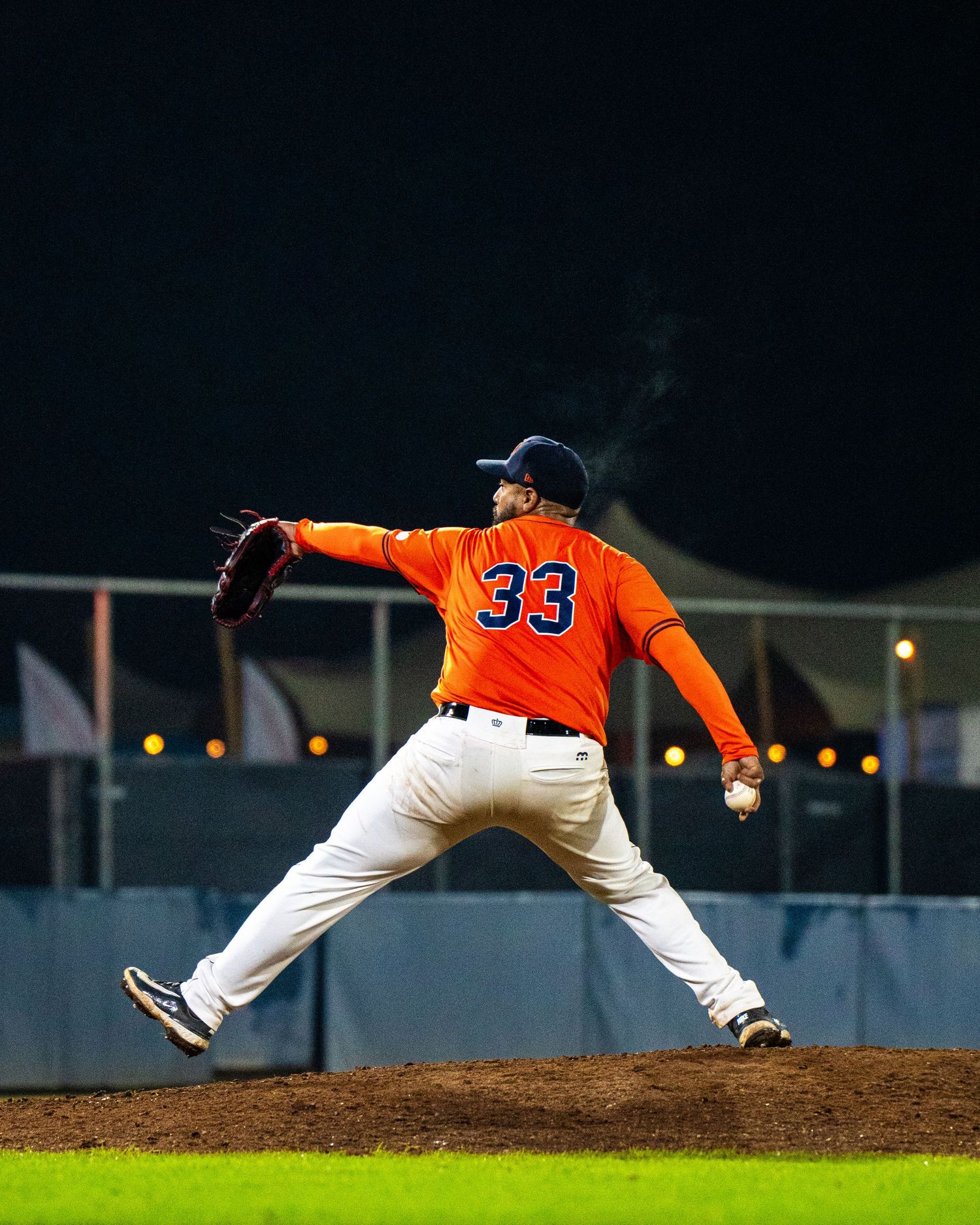 Baseball pitcher in orange jersey number 33 follows through on delivery motion during nighttime game.