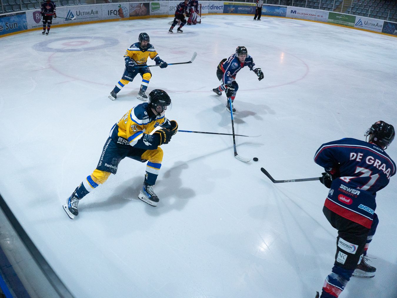 Ice hockey players in yellow and blue jerseys compete for the puck during an intense match on the rink.