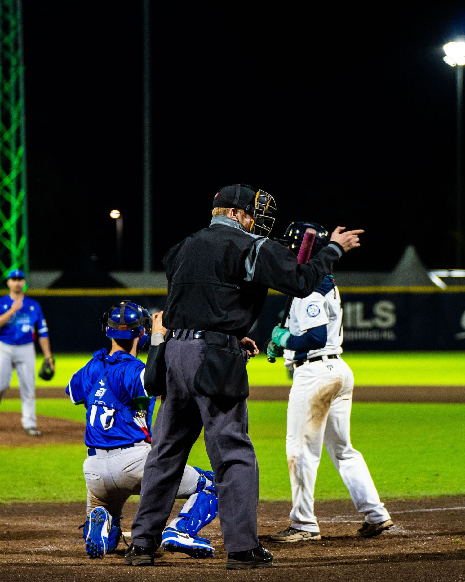 Baseball umpire in black uniform gestures during a night game with players in blue and white uniforms visible.