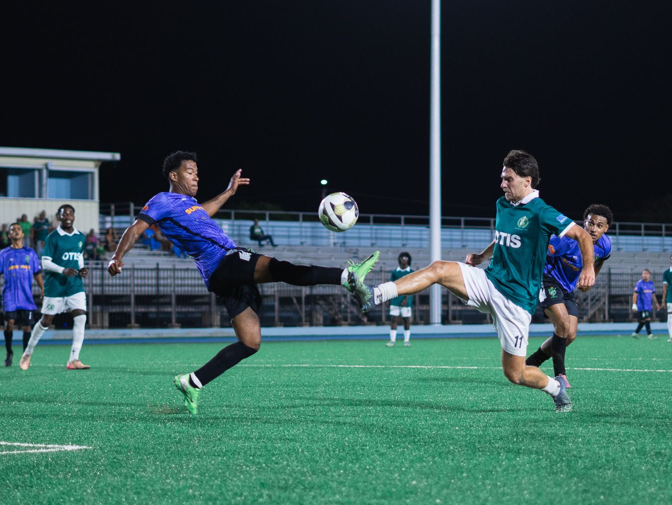 Soccer players compete for the ball on a green turf field during a night game in blue and green kits.