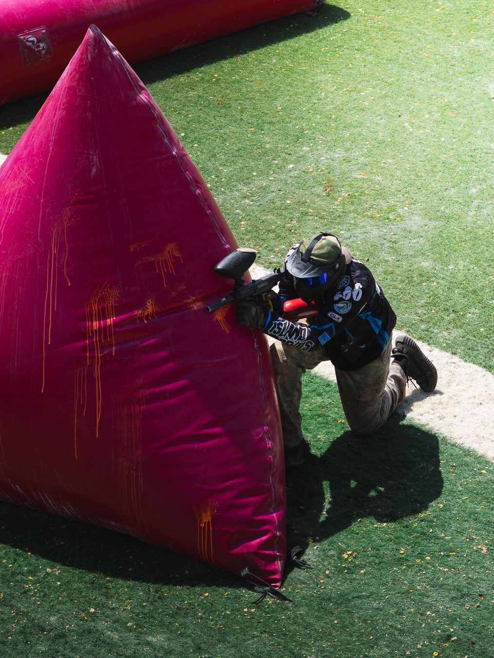 A paintball player in protective gear crouches behind a large red inflatable bunker on a green grass field.