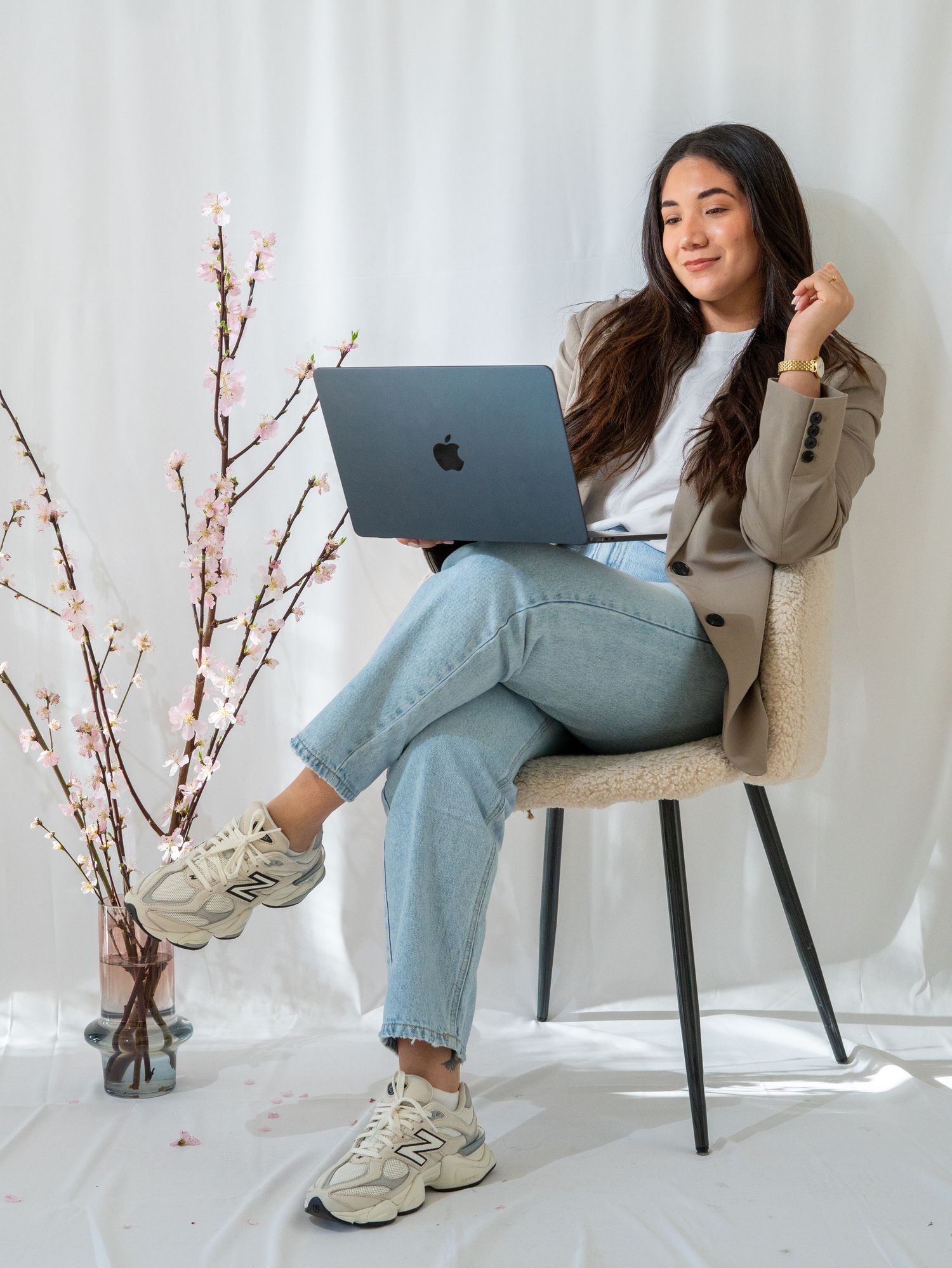 Person in beige coat and light jeans sits casually on modern chair with laptop while cherry blossoms decorate scene.