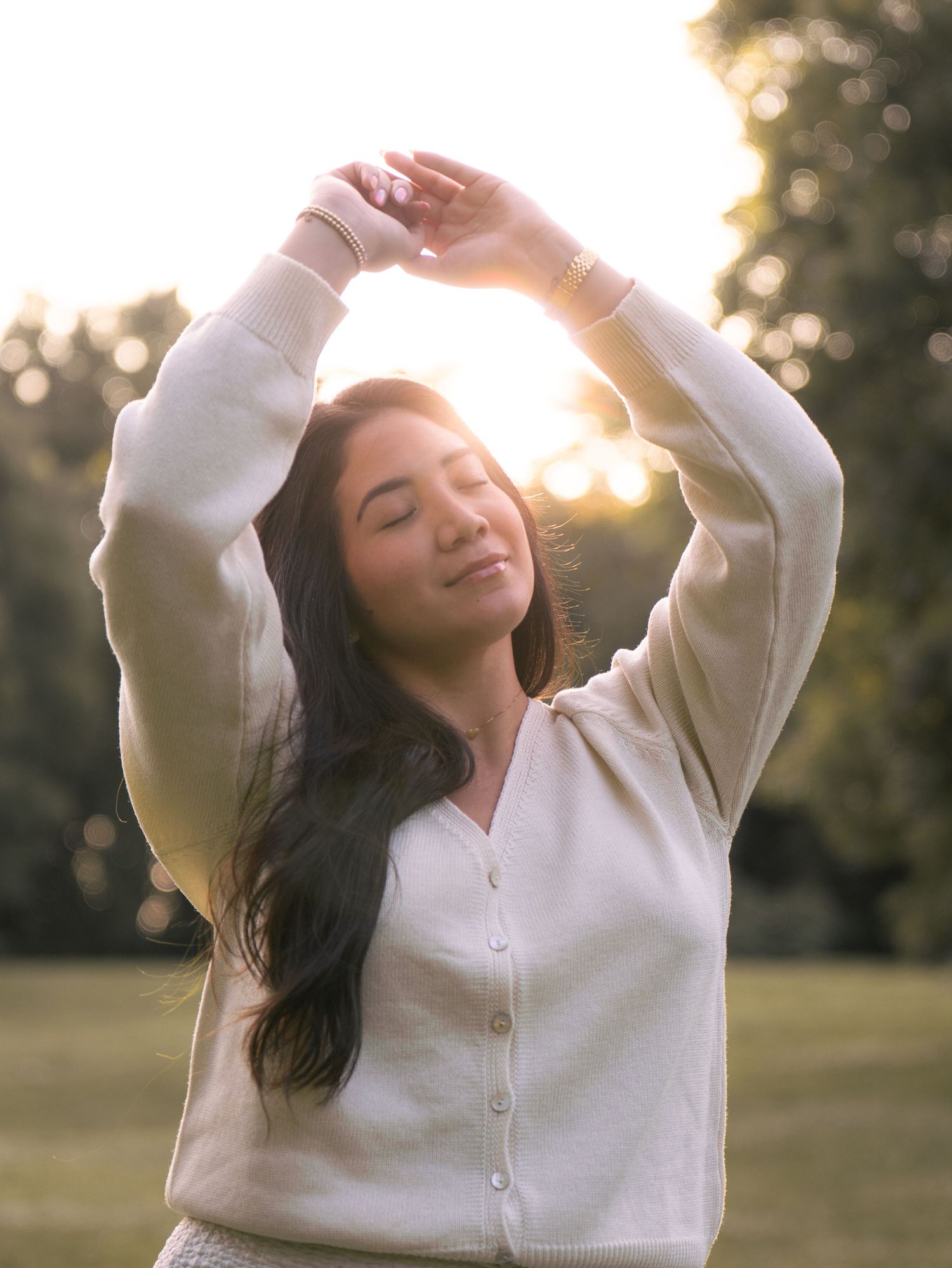 Person in white sweater dancing joyfully in a sunlit park during golden hour with flowing dark hair.