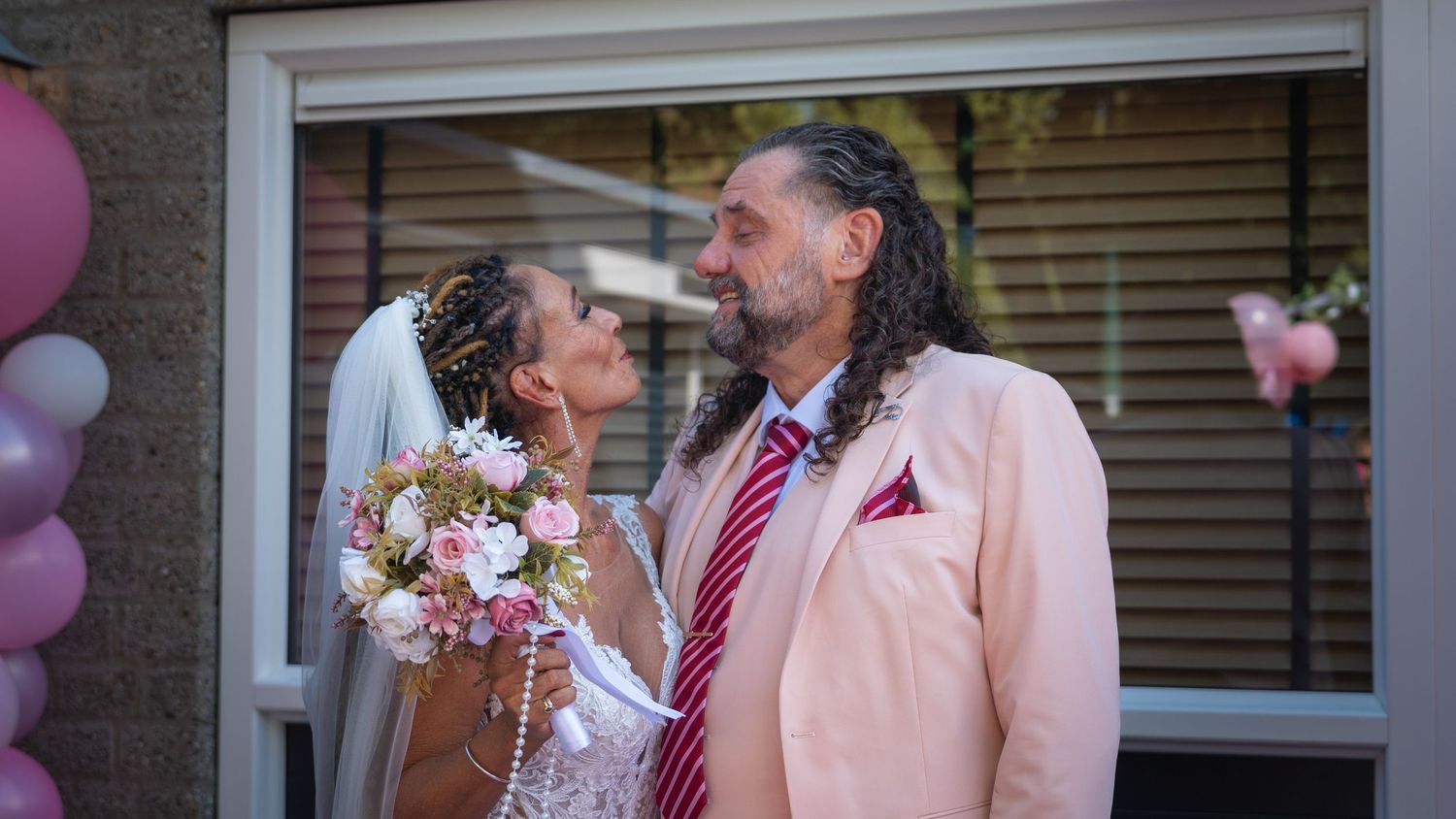 A happy couple shares a joyful moment at their wedding ceremony with pink and purple balloons in the background.