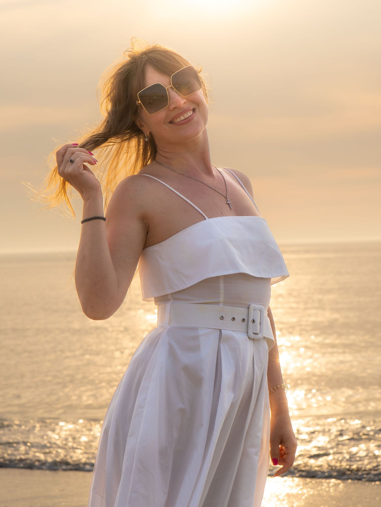 A woman in a flowing white dress twirls joyfully on the beach at sunset with the ocean sparkling behind her.