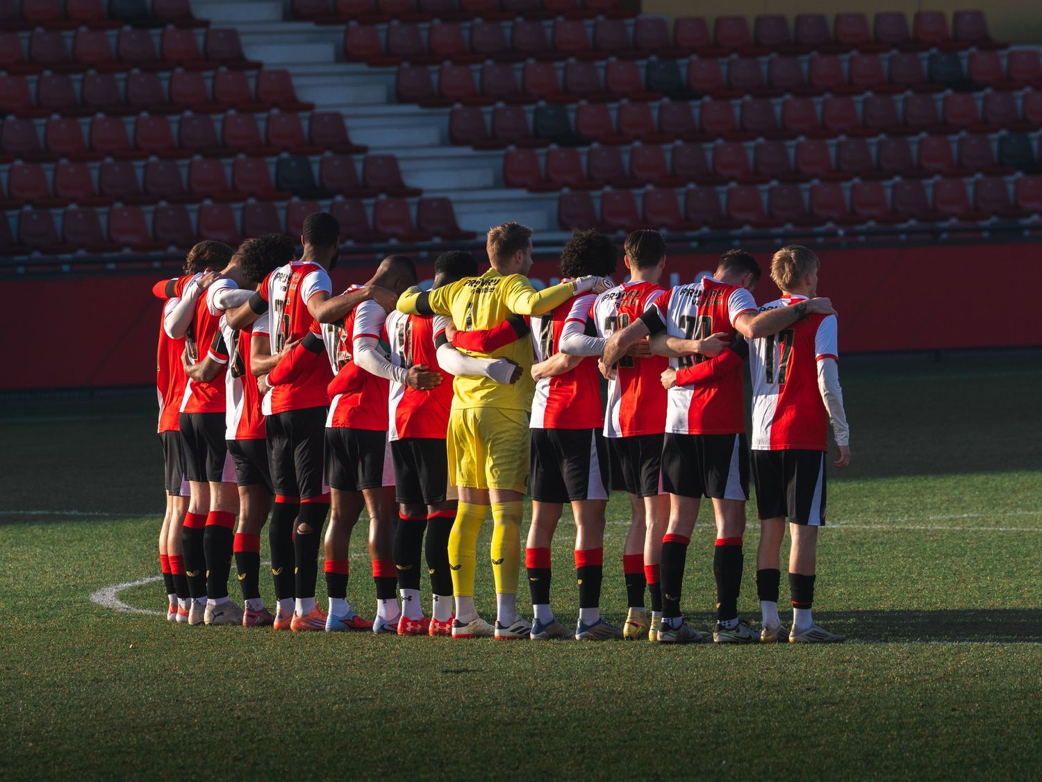 Soccer team huddles together on field wearing red and white jerseys with one player in yellow goalkeeper uniform.