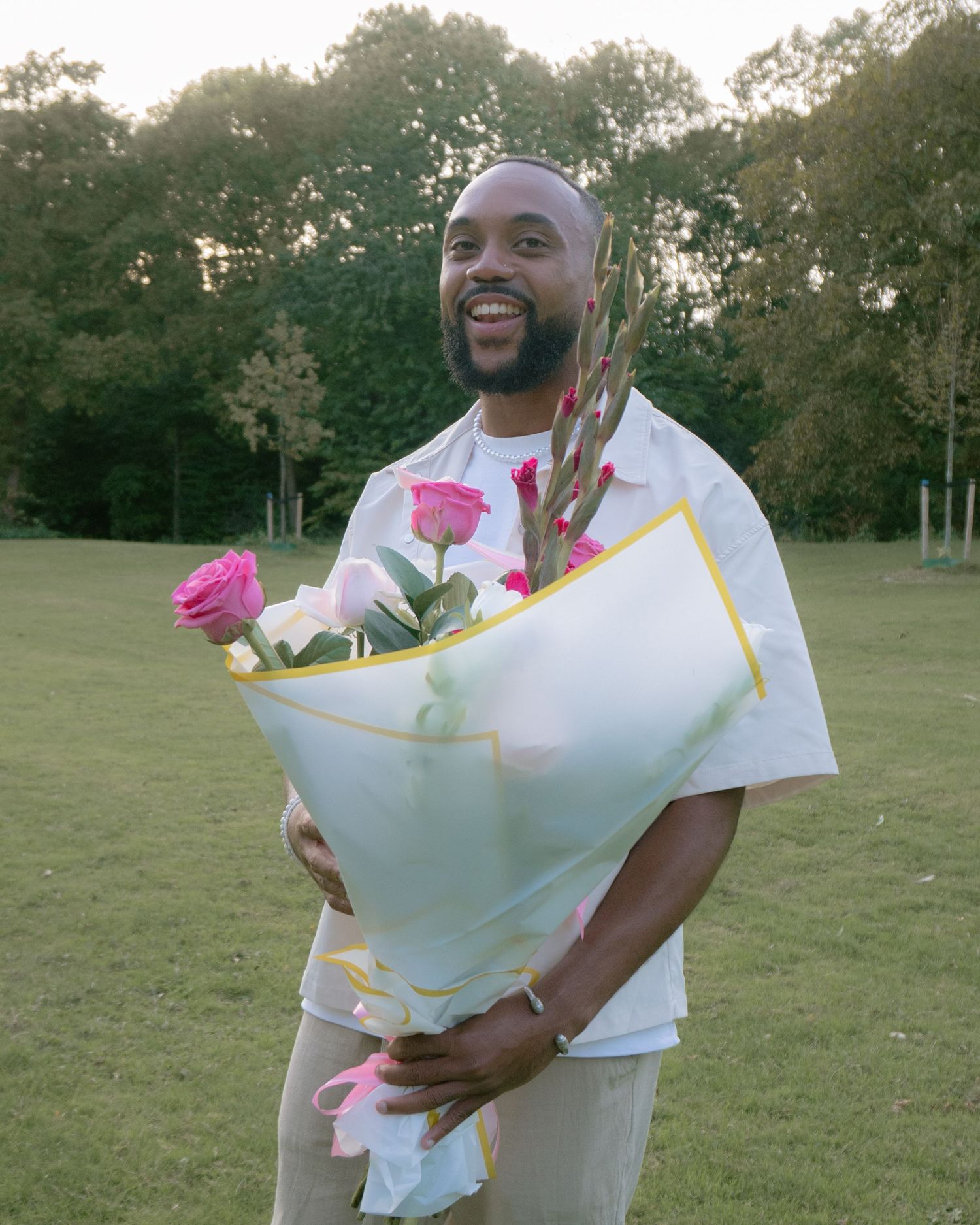 Someone in a white outfit stands outdoors on a grassy field holding a large bouquet wrapped in light blue paper.