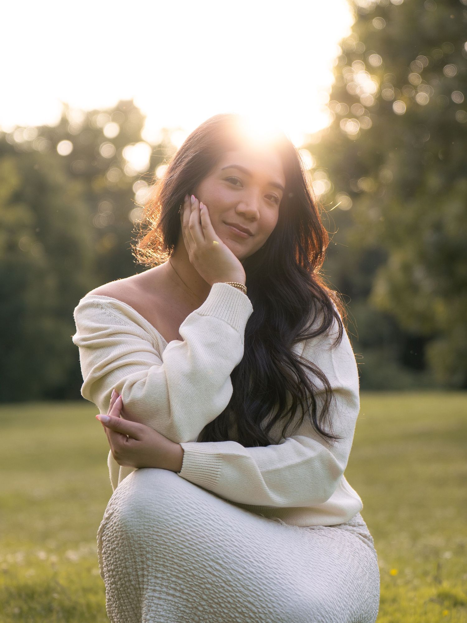 A young woman in a cream off-shoulder sweater poses gracefully outdoors at golden hour with sunlight glowing behind her.