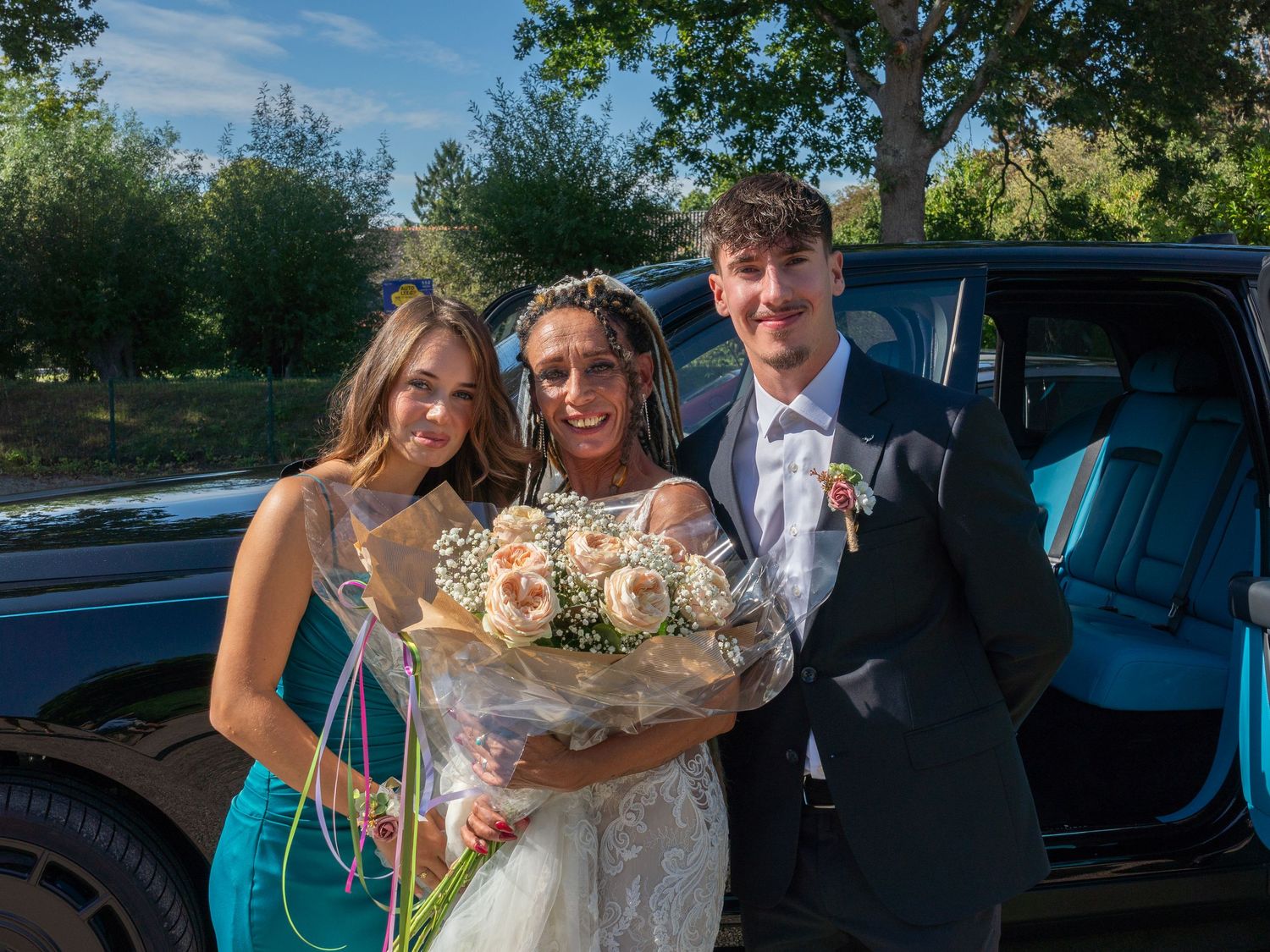 A group of wedding attendees gather around vehicles in an outdoor parking area on a sunny day.