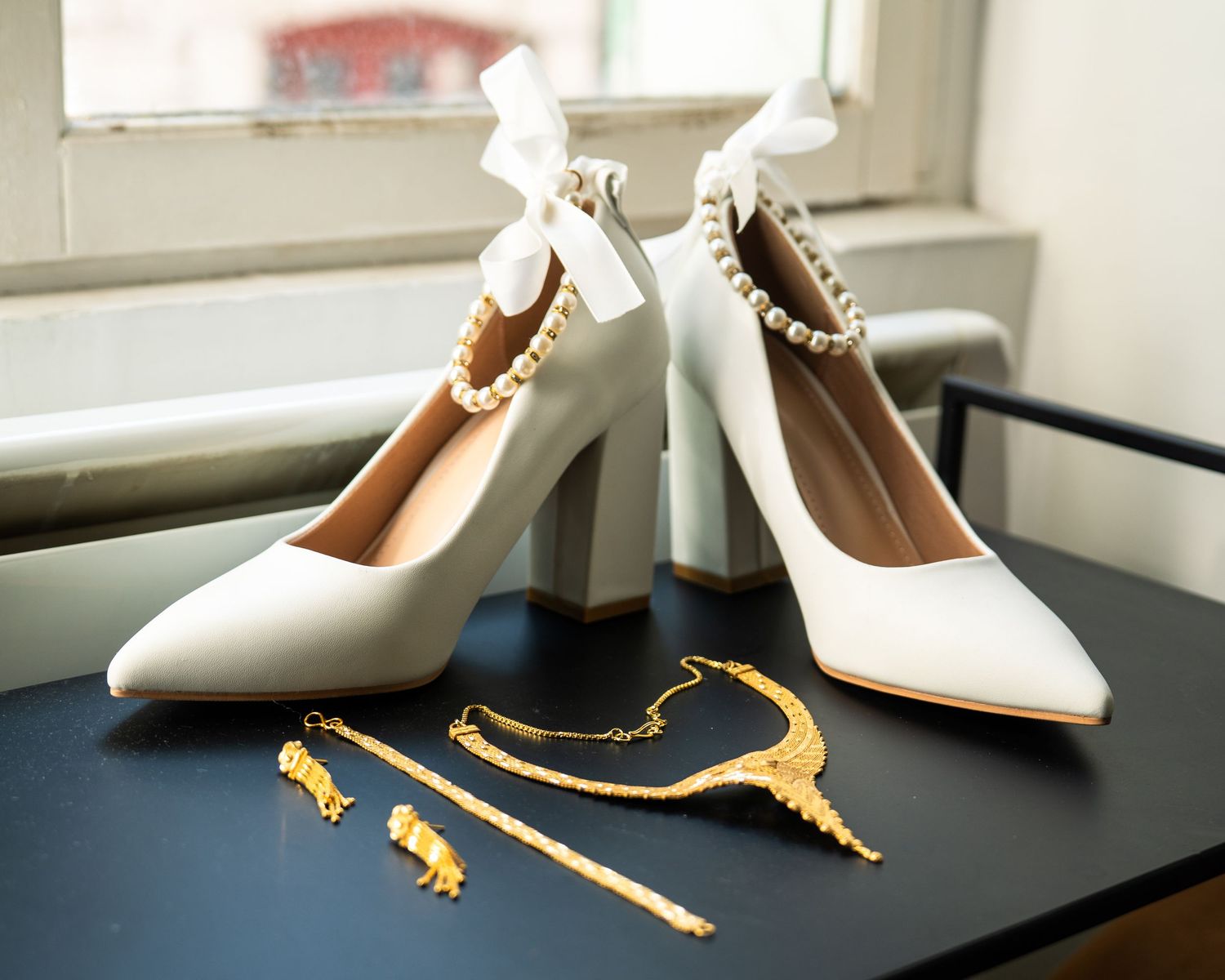White leather high heels with gold hardware details sit on a dark surface near a window.