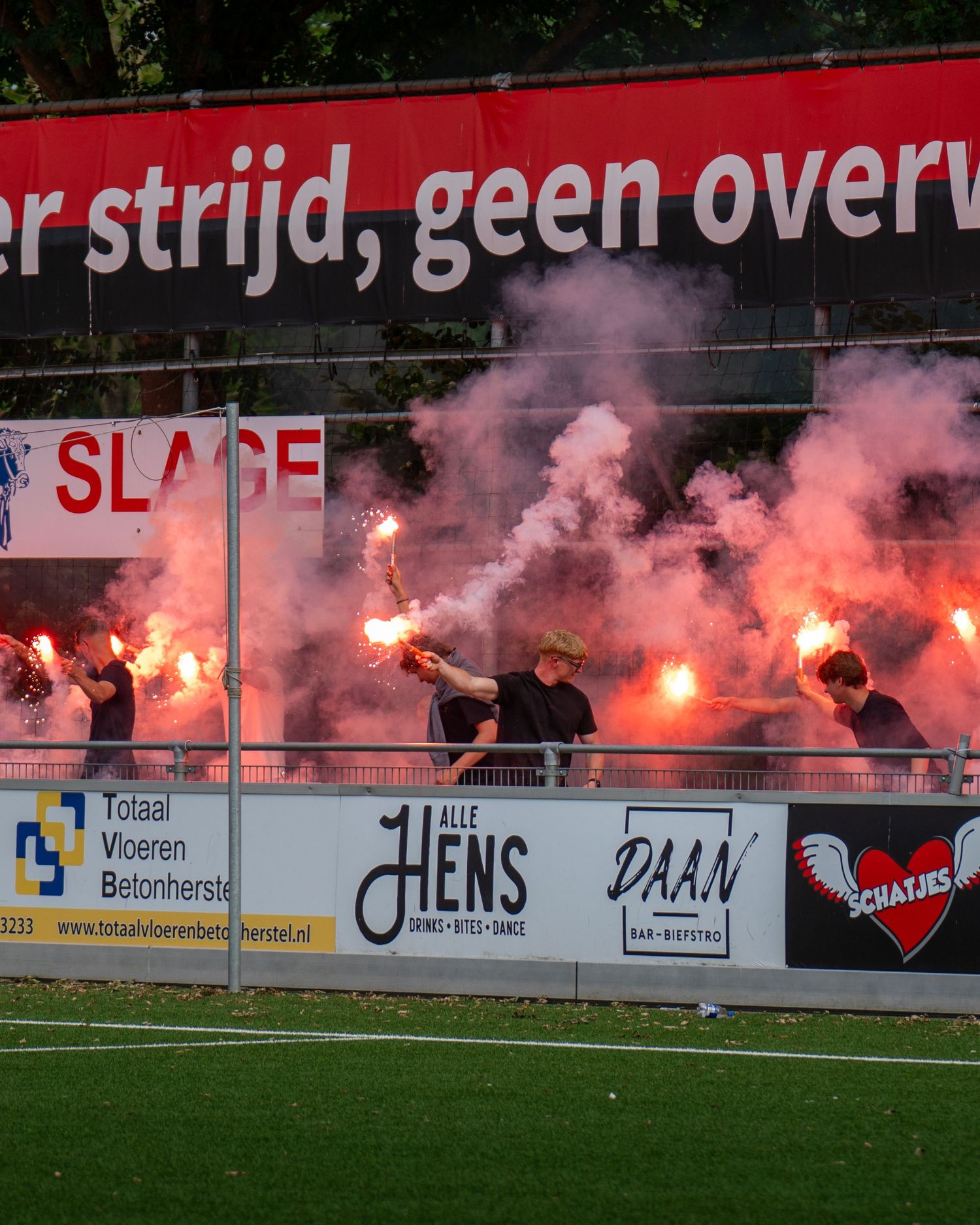 Red flares and smoke illuminate a soccer stadium as supporters create an intense atmosphere during a match.