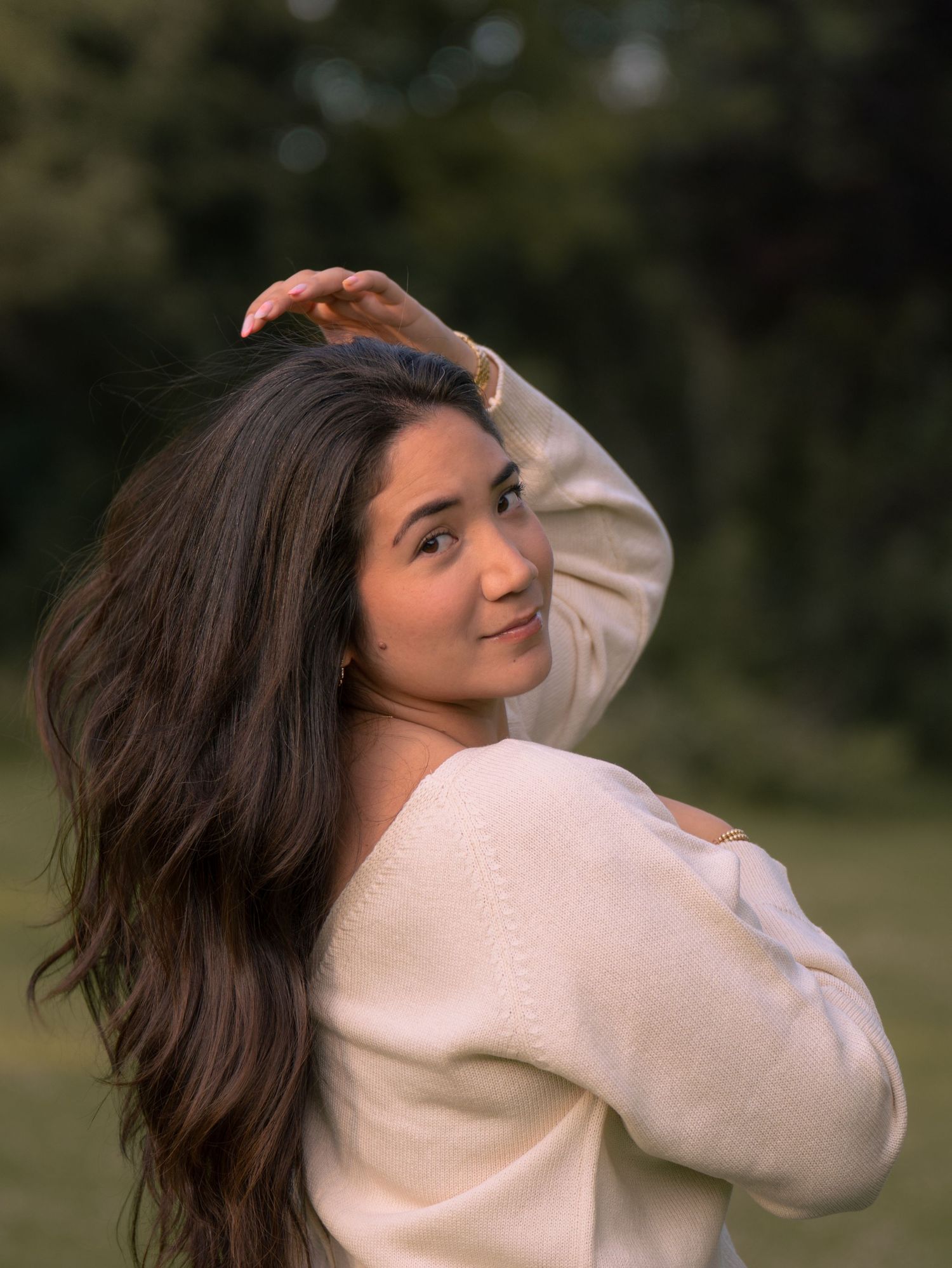 Portrait of someone in a cream sweater looking over their shoulder with long dark hair flowing in an outdoor setting.