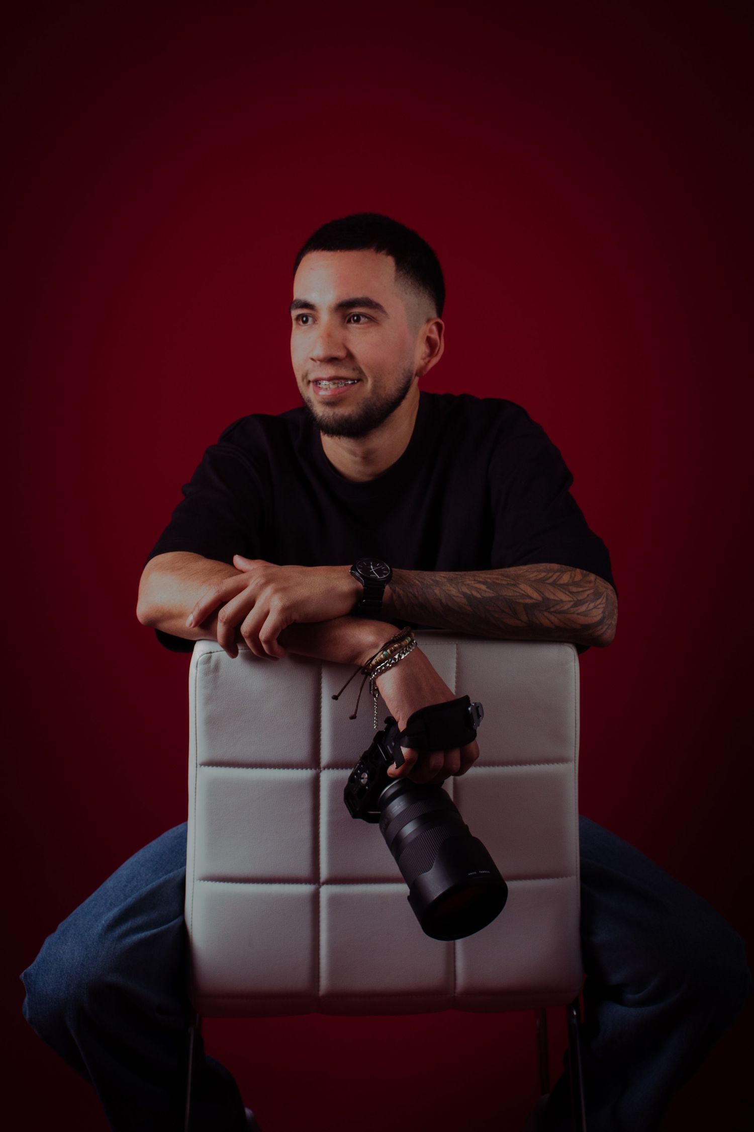 Professional photographer wearing dark clothing sits on white couch with camera against deep red backdrop.