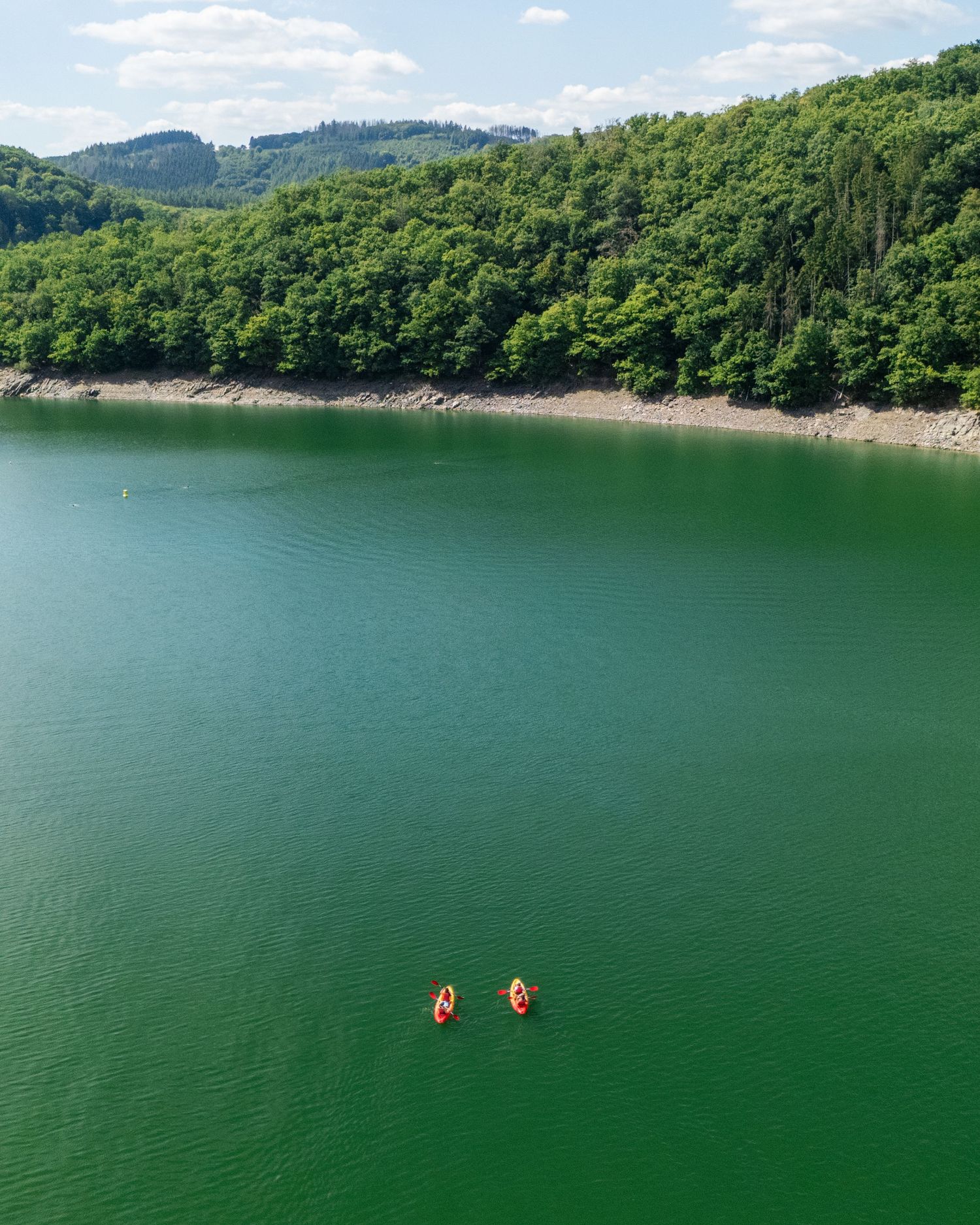 Aerial view of two kayakers paddling on a serene green lake surrounded by lush forested hills.