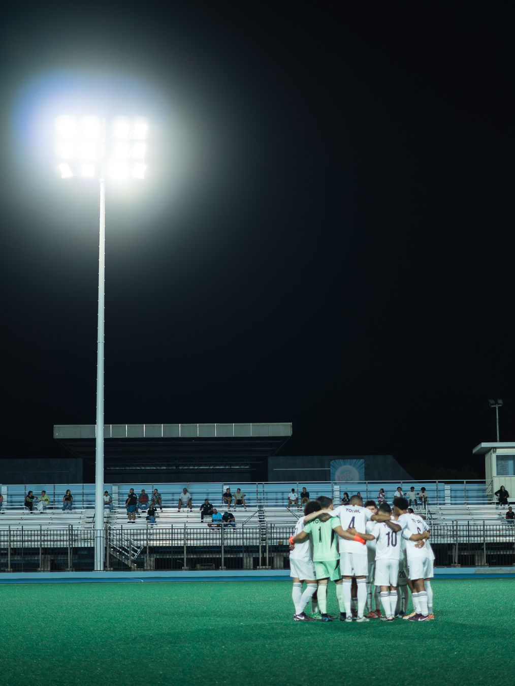 Soccer team huddles under bright stadium lights on a green field at night with bleachers in background.