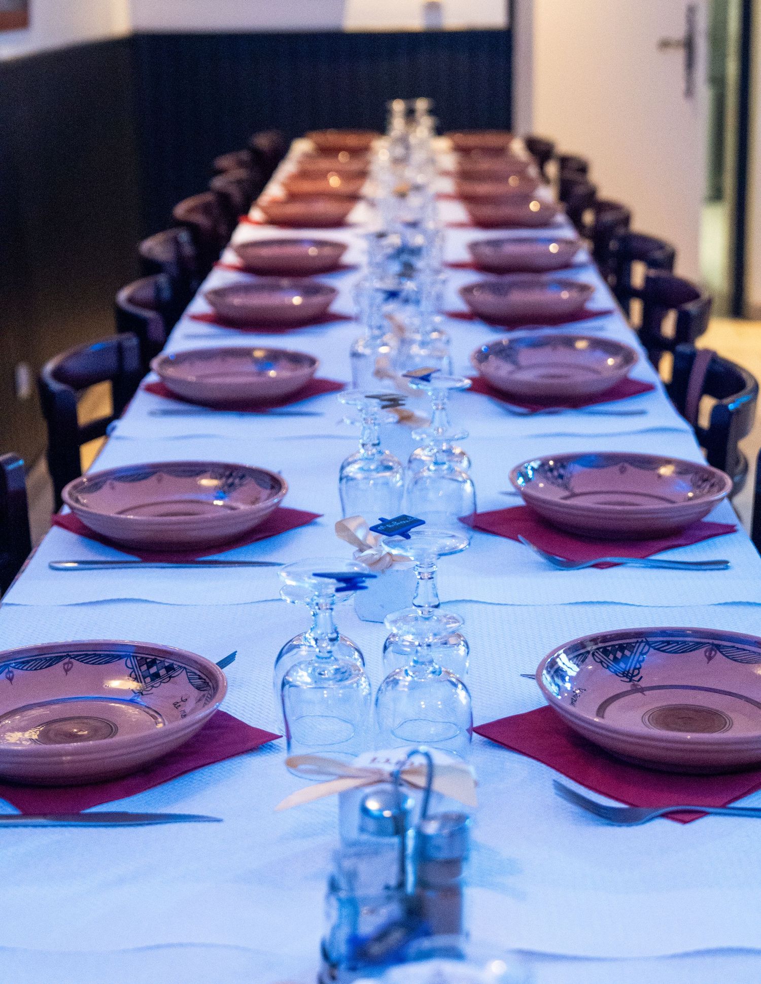 A long formal dining table set with burgundy plates and water glasses illuminated by cool blue lighting.
