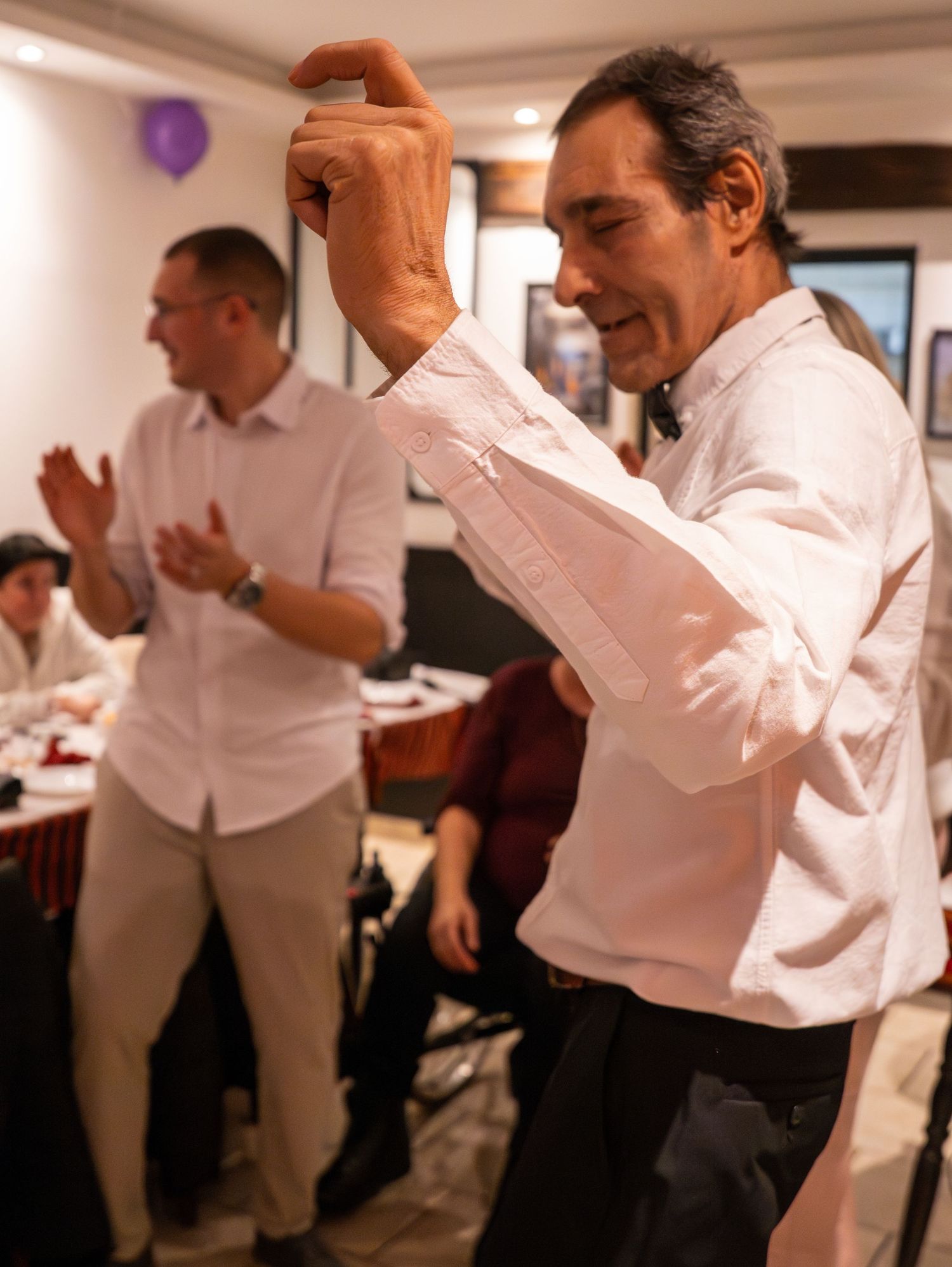 A person in formal white attire dances enthusiastically at an indoor party with purple decorations.