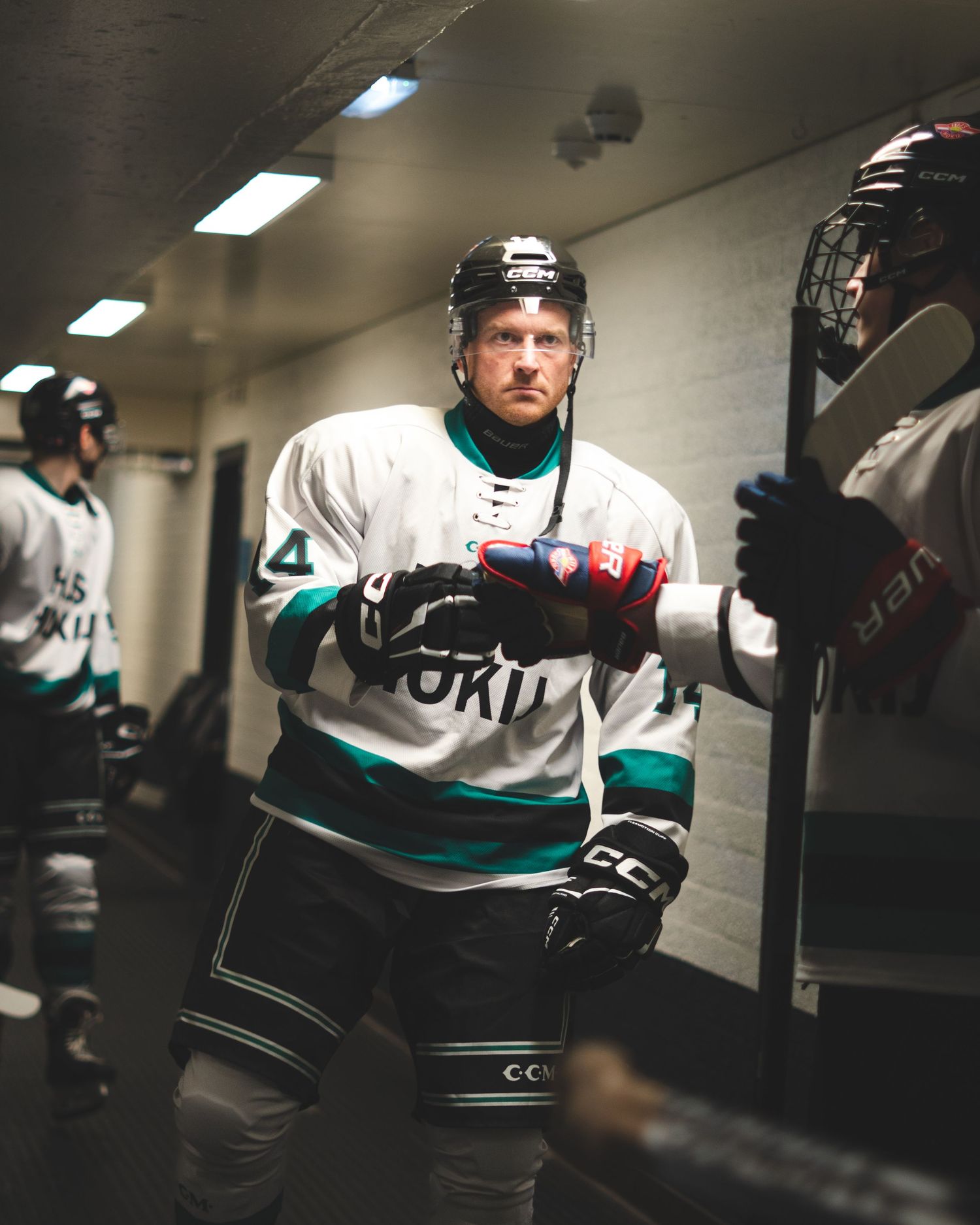 Hockey players in white and teal jerseys gather in a dimly lit locker room hallway before a game.