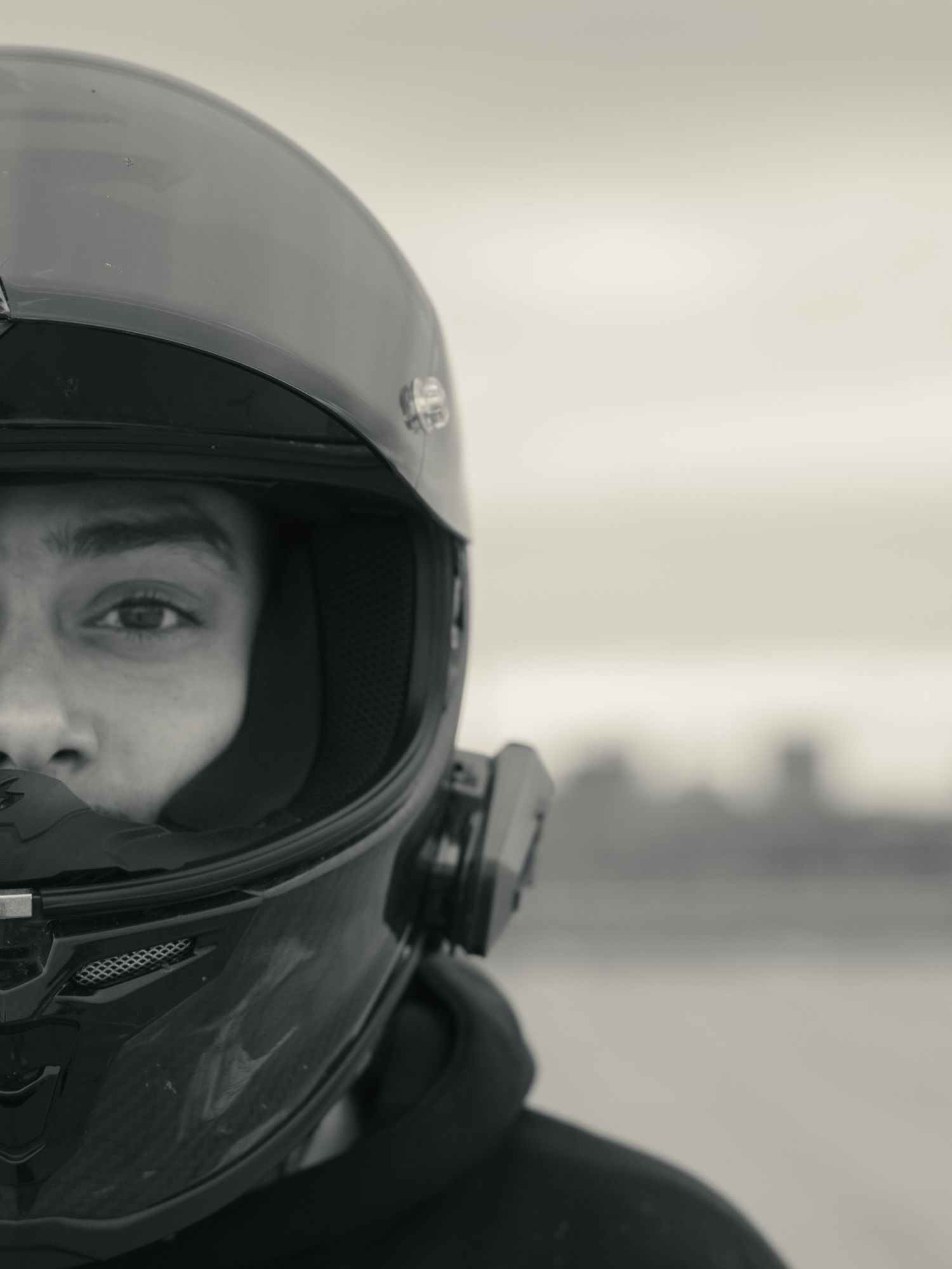 A close-up black and white portrait of a motorcyclist wearing a dark helmet against an overcast sky.