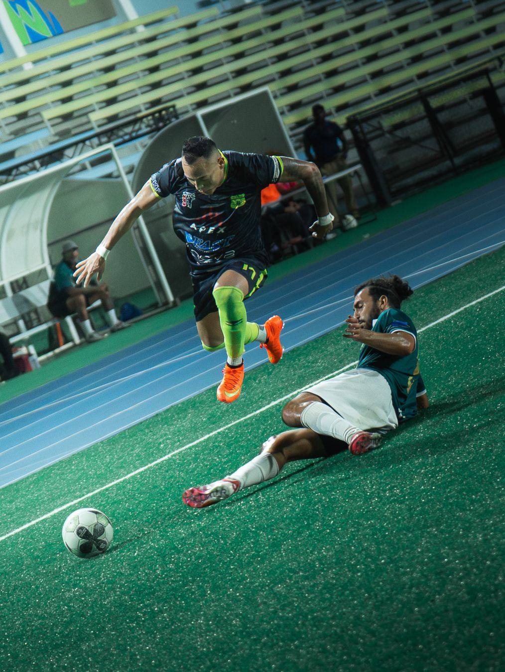 A soccer player leaps over a sliding tackle during an indoor futsal match on a green court.
