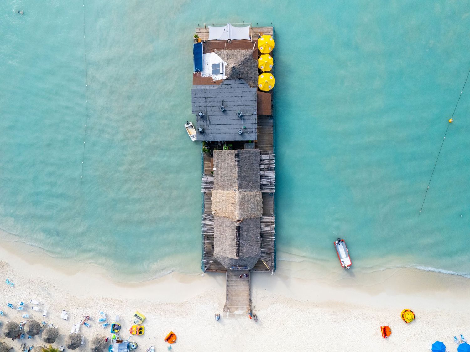 Aerial view of a wooden pier and beach bar extending into turquoise Caribbean waters with white sandy beach.