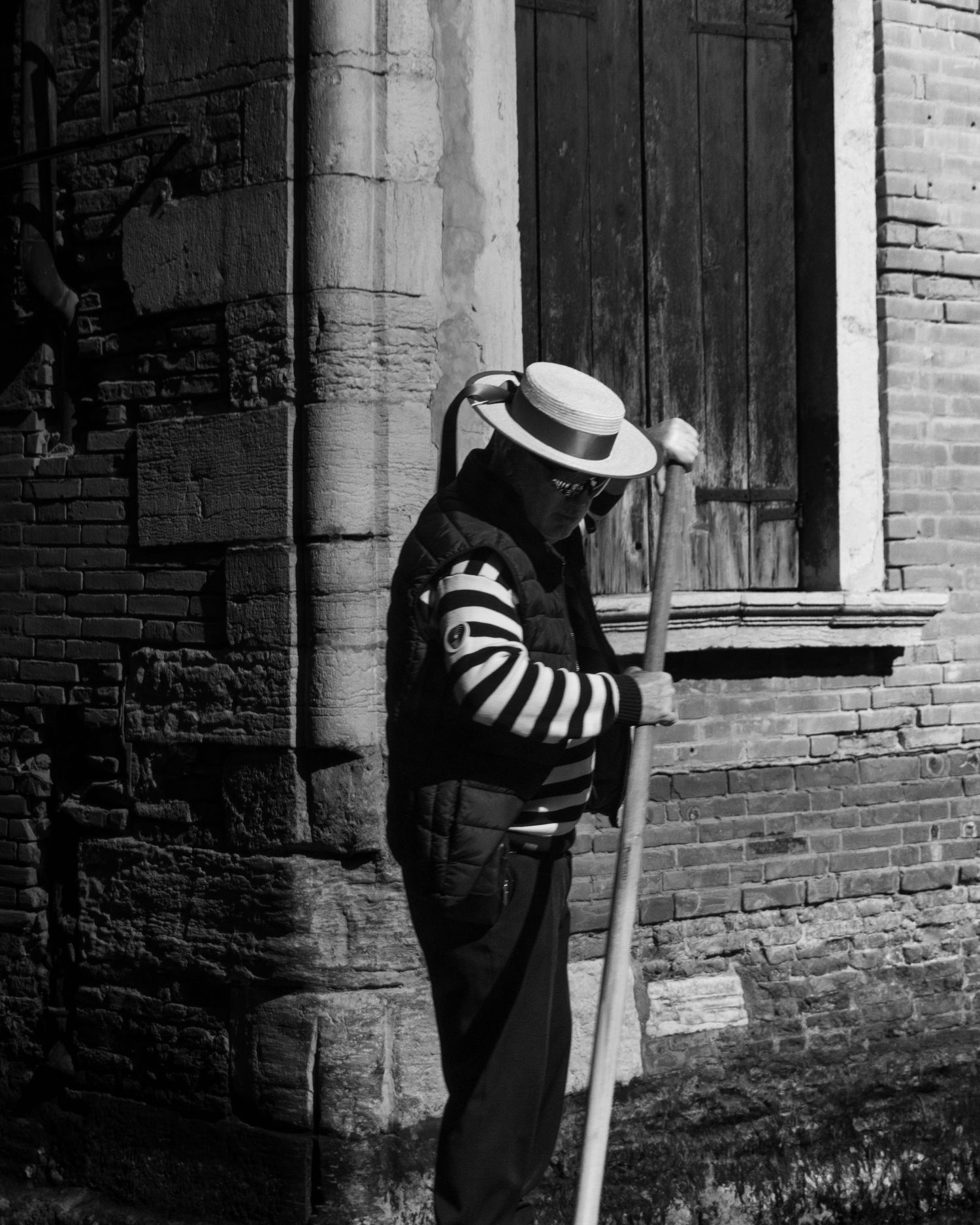 Black and white photo of a gondolier in striped shirt and straw hat leaning on an oar beside a brick building in Venice.