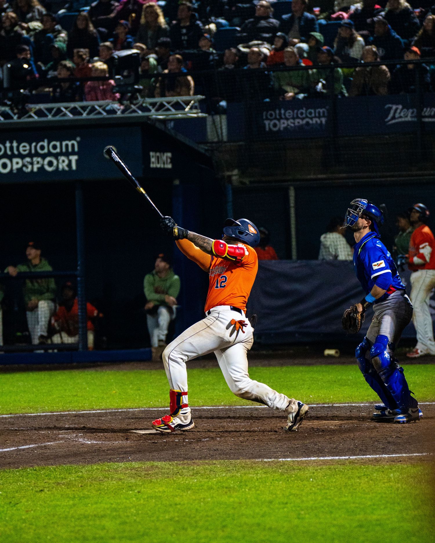 Baseball batter in orange uniform takes a powerful swing at home plate during nighttime game.