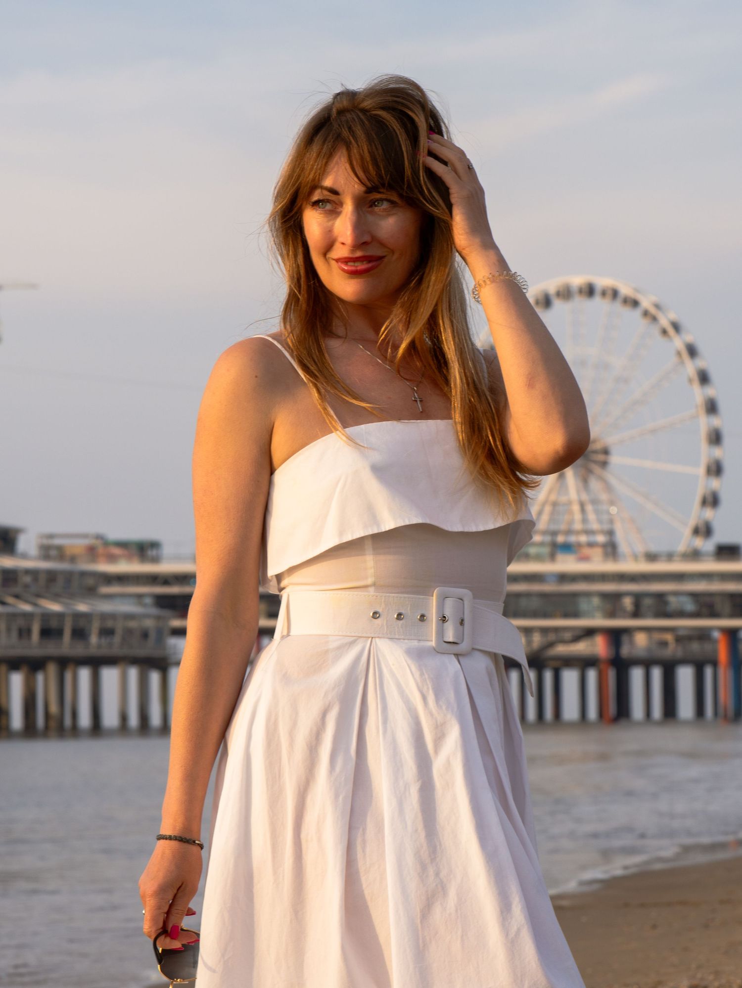 A woman in a white sundress poses on the beach with a Ferris wheel visible in the background during golden hour.