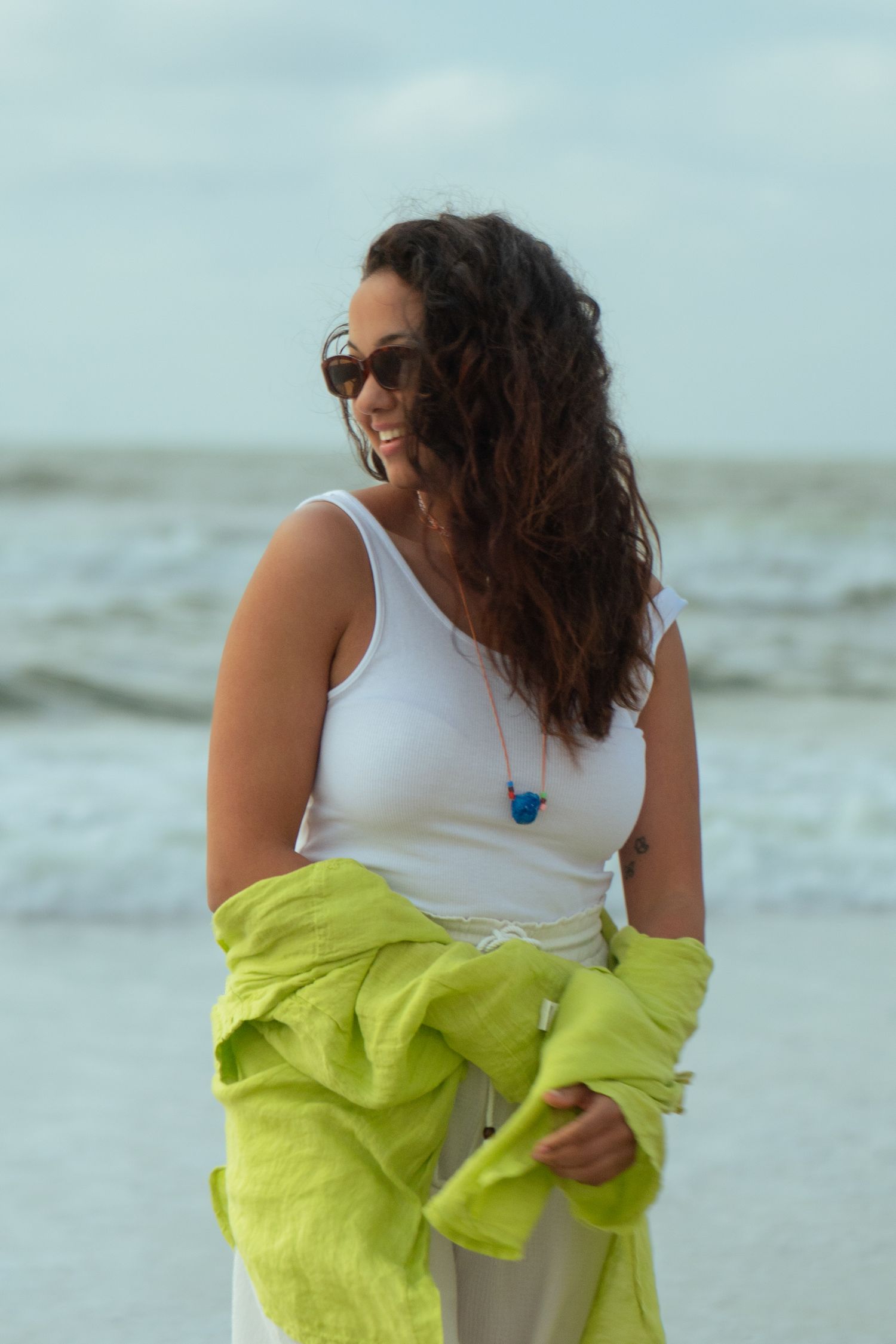 Someone with long curly hair wearing sunglasses and a white tank top holds a lime green jacket at the beach.