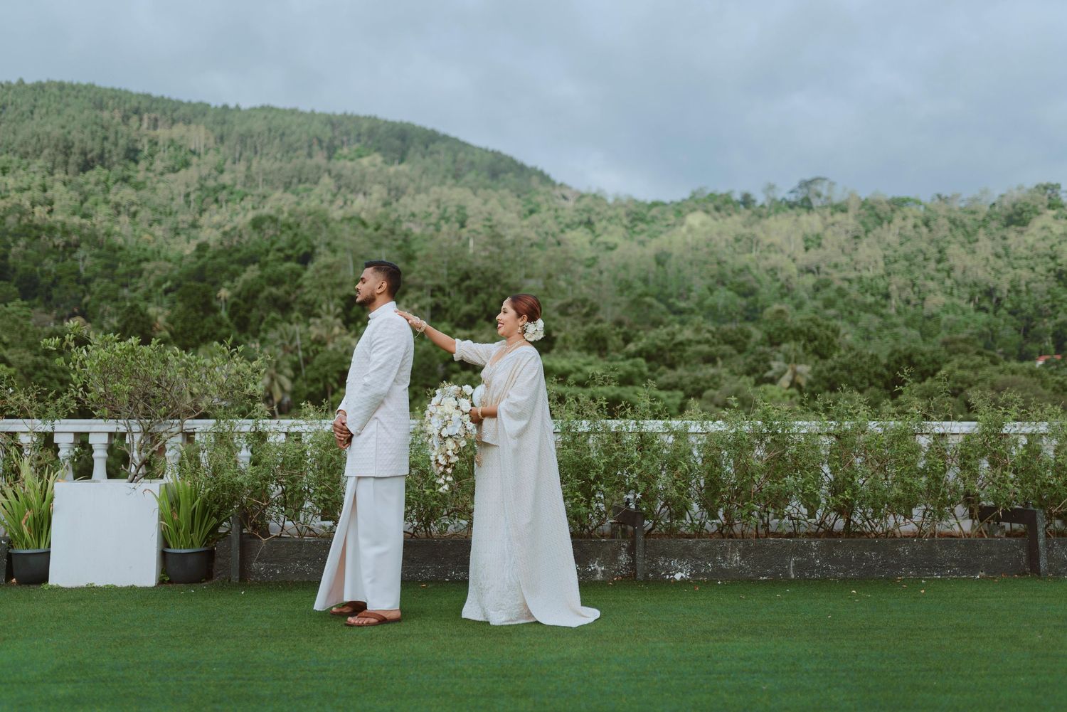 A couple in white wedding attire stands together on a lawn terrace overlooking lush green hills and forest.