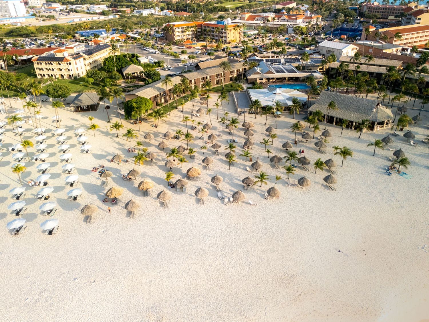 Aerial view of a tropical beach resort with thatched umbrellas, white sand, palm trees, and colorful buildings.