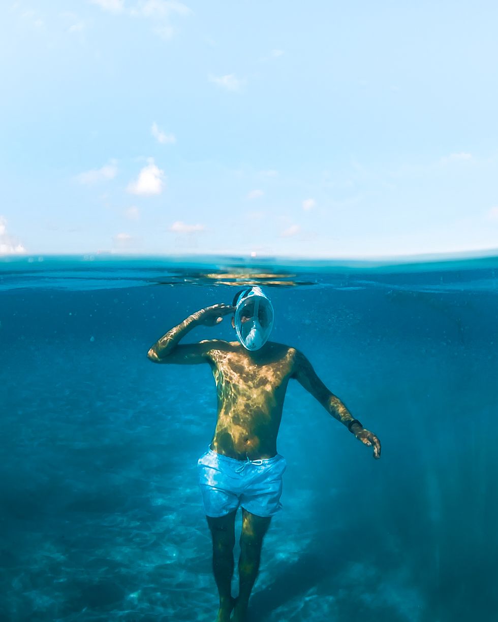 Shirtless snorkeler wearing full-face mask stands waist-deep in clear tropical blue ocean, split underwater view.