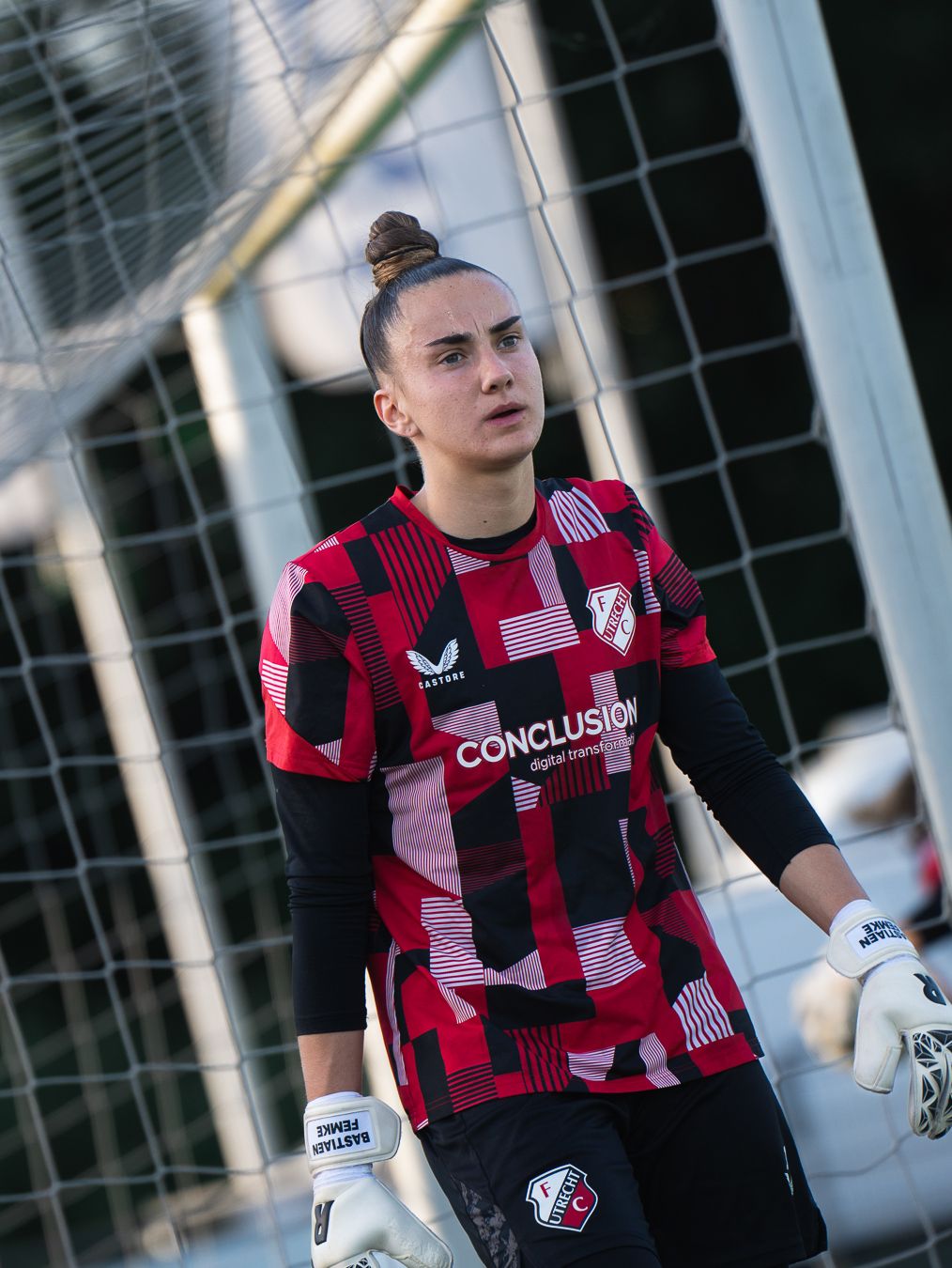 Female goalkeeper in red and black Castore kit with Conclusion sponsor stands focused near goal post.