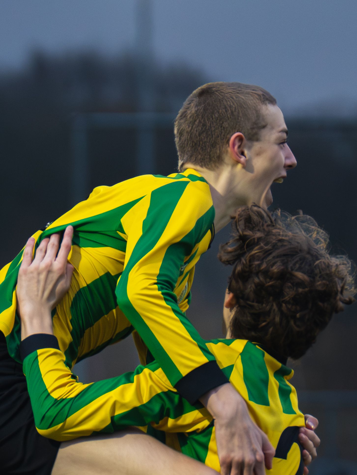 Two soccer players in yellow and green striped jerseys embrace in celebration during a match.