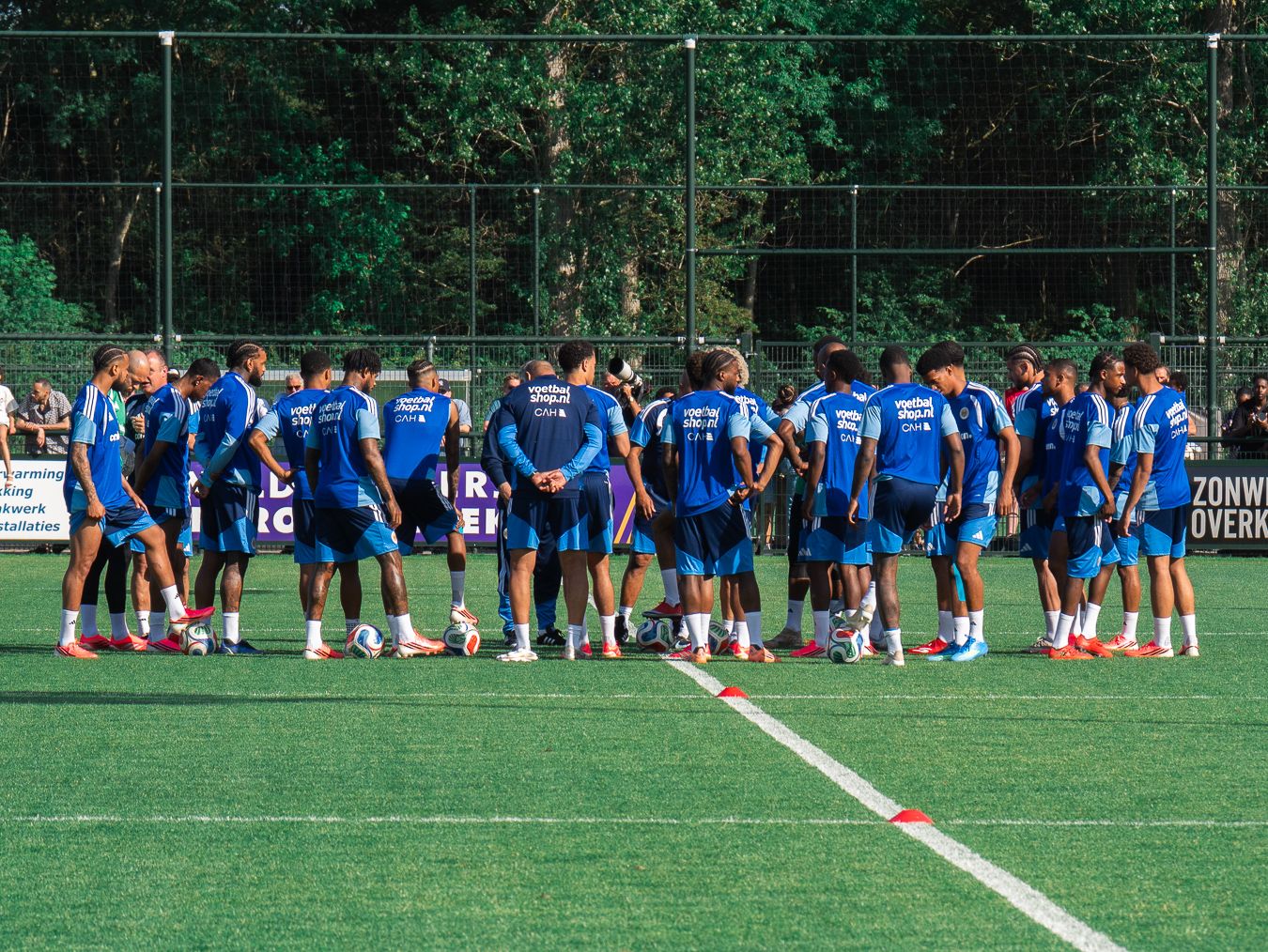 Soccer team in blue uniforms huddles on a green turf field during a training session surrounded by trees.