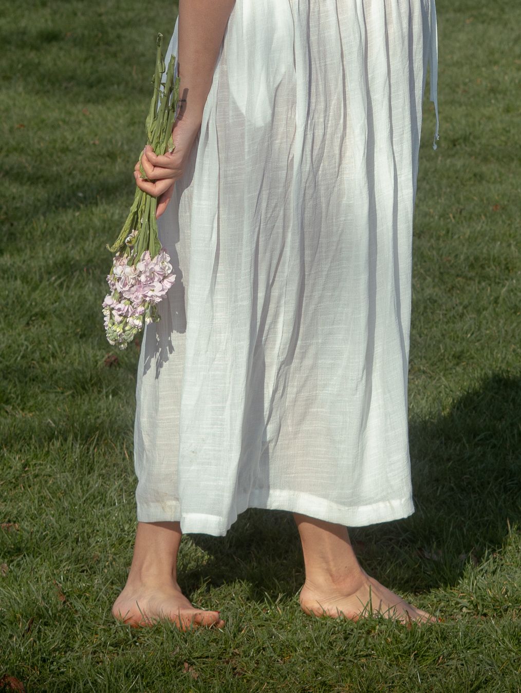 Barefoot woman in white linen dress holding purple wildflowers while standing on green grass.