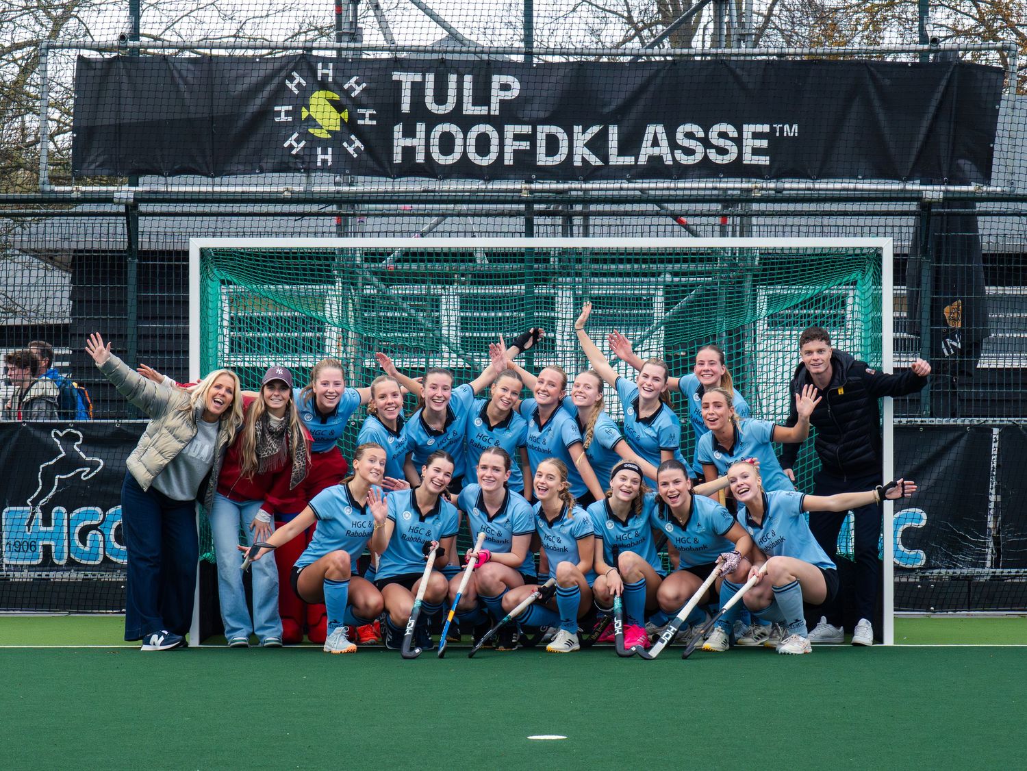 Field hockey team celebrates together on turf field beneath Tulp Hoofdklasse banner at sports facility.