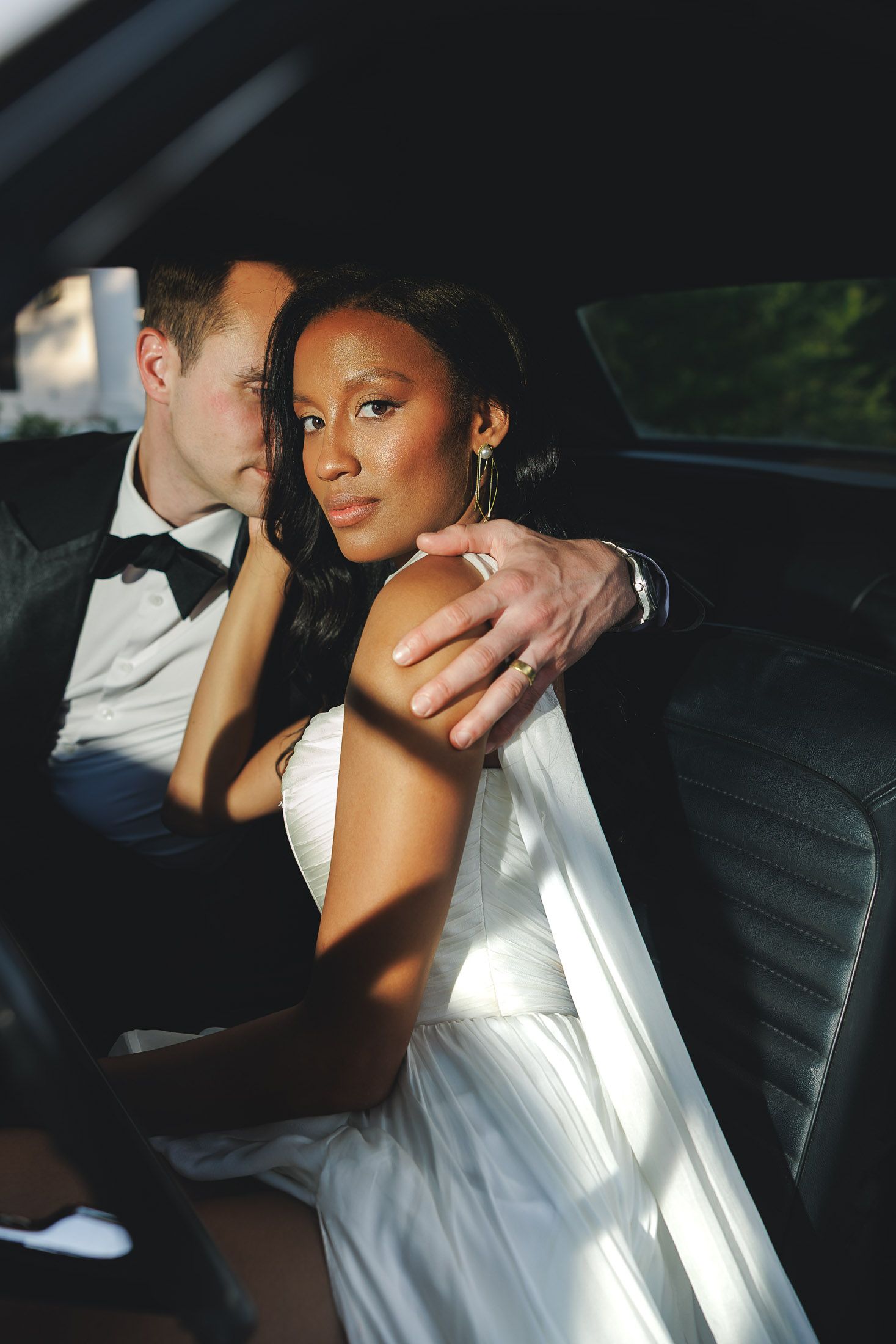 Elegant interracial couple in wedding attire sitting in a car, bride in white gown with diamond ring, groom in black tuxedo.